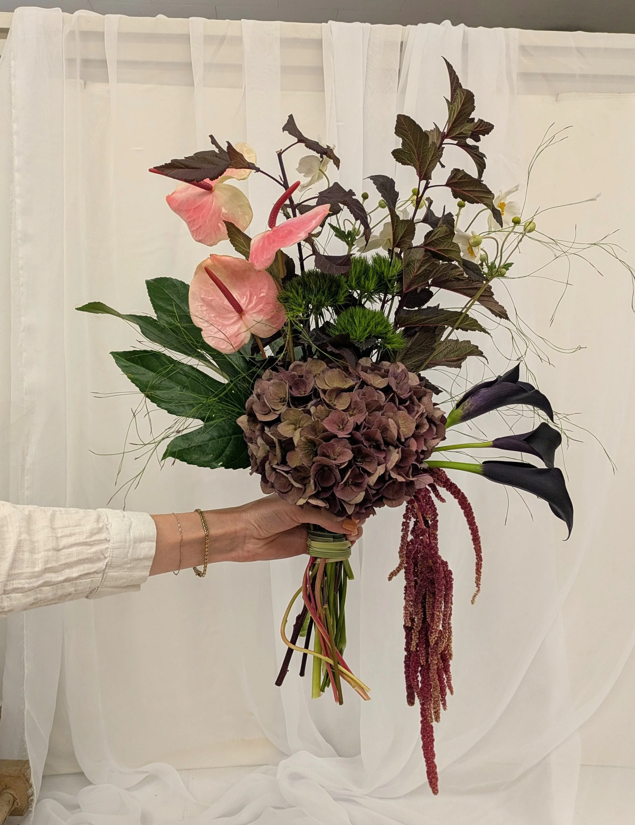 Person holding a bouquet of various flowers including pink anthuriums, dark purple calla lilies, hydrangea, green chrysanthemums, and amaranthus, in front of a light-colored fabric backdrop.