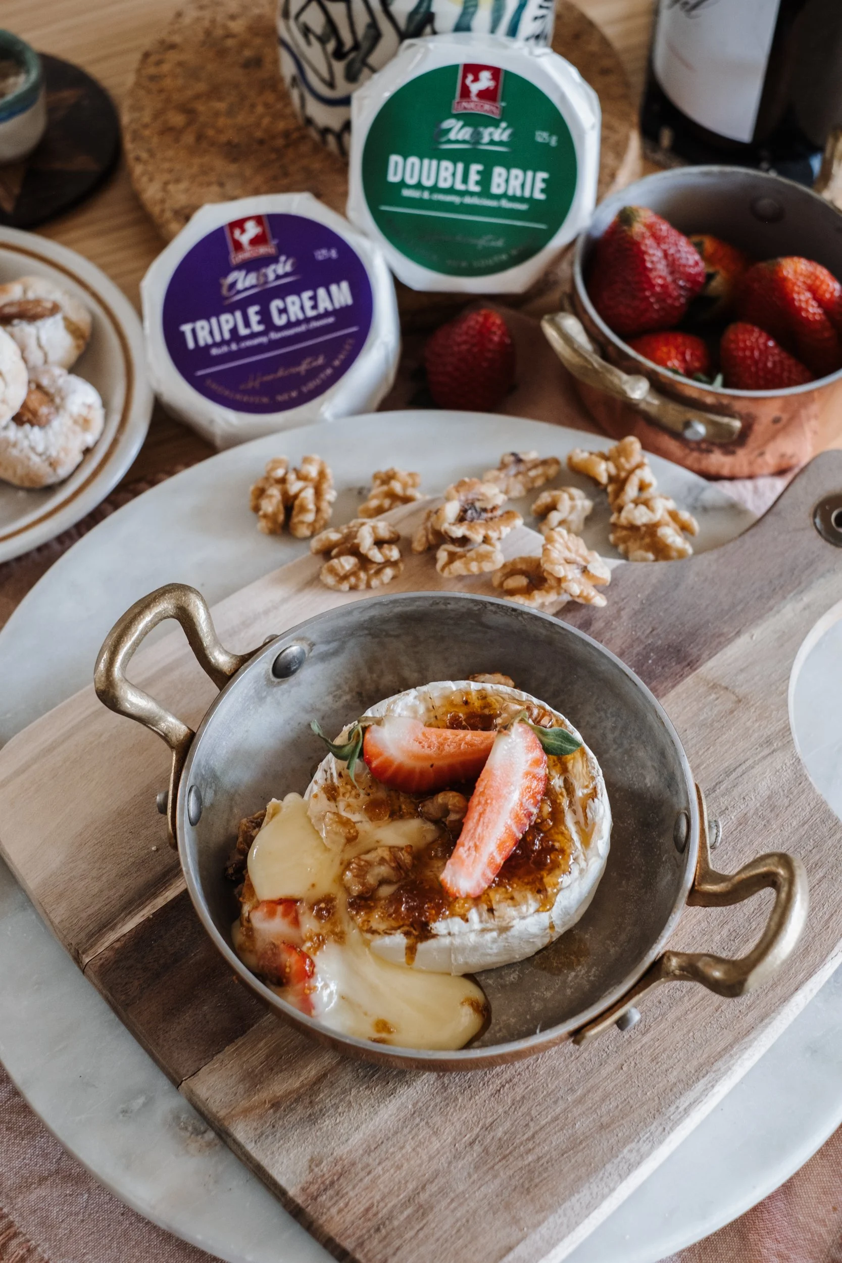 A gooey baked brie with fresh strawberries, fig jam and crumbled walnuts. The gooey cheese is oozing out into the rustic copper pan. Unicorn Cheese Triple Cream brie is in the background.