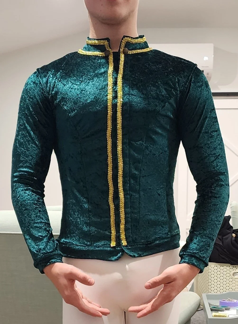 Person wearing a dark green velvet jacket with gold trim, standing indoors.