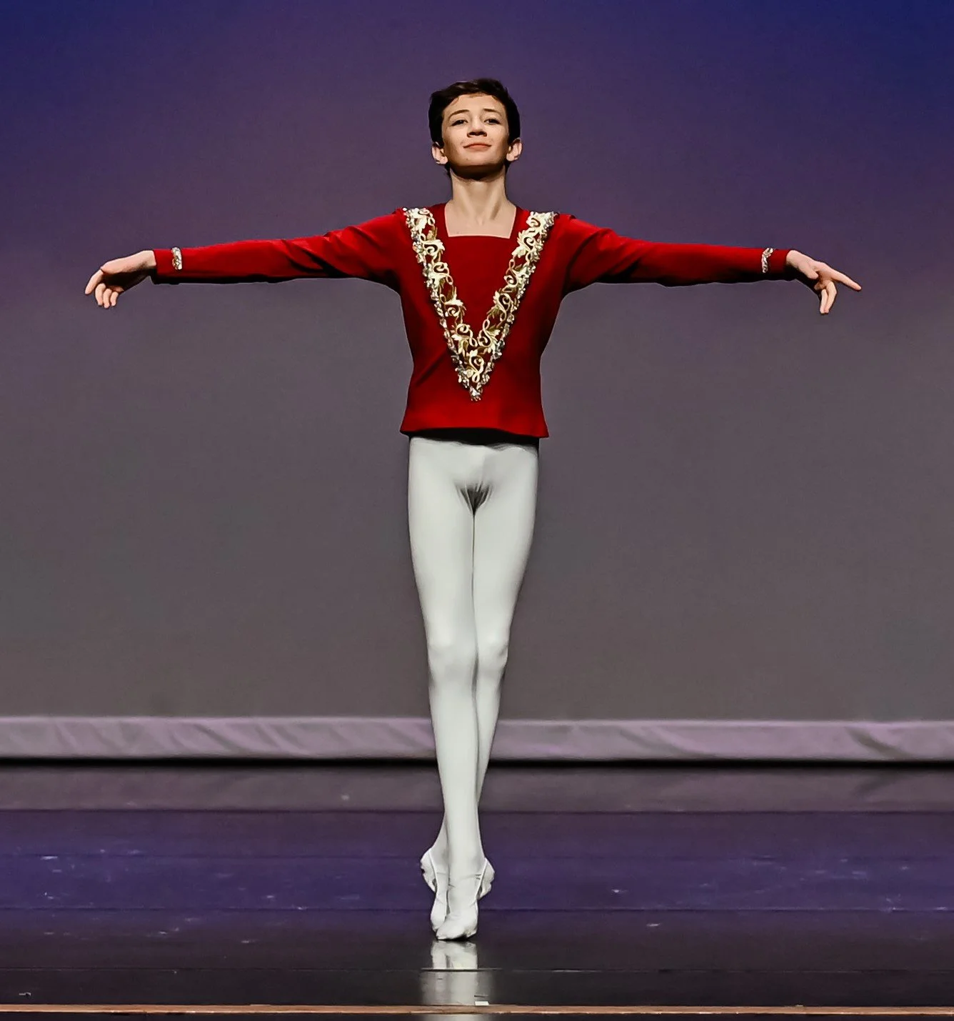A young ballet dancer standing in a ballet pose with arms extended outward and one leg slightly crossed in front of the other on a stage, wearing a red costume with gold embroidery and white tights.