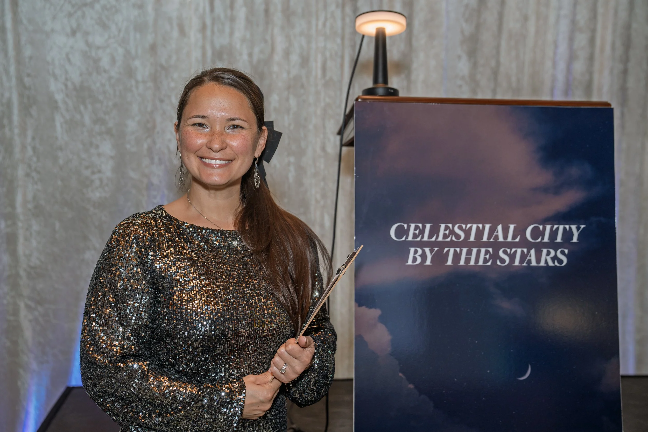 A woman with long brown hair wearing a black and gold sequined dress, smiling, holding a clipboard, standing beside a sign that reads 'Celestial City by the Stars' with a picture of the night sky.