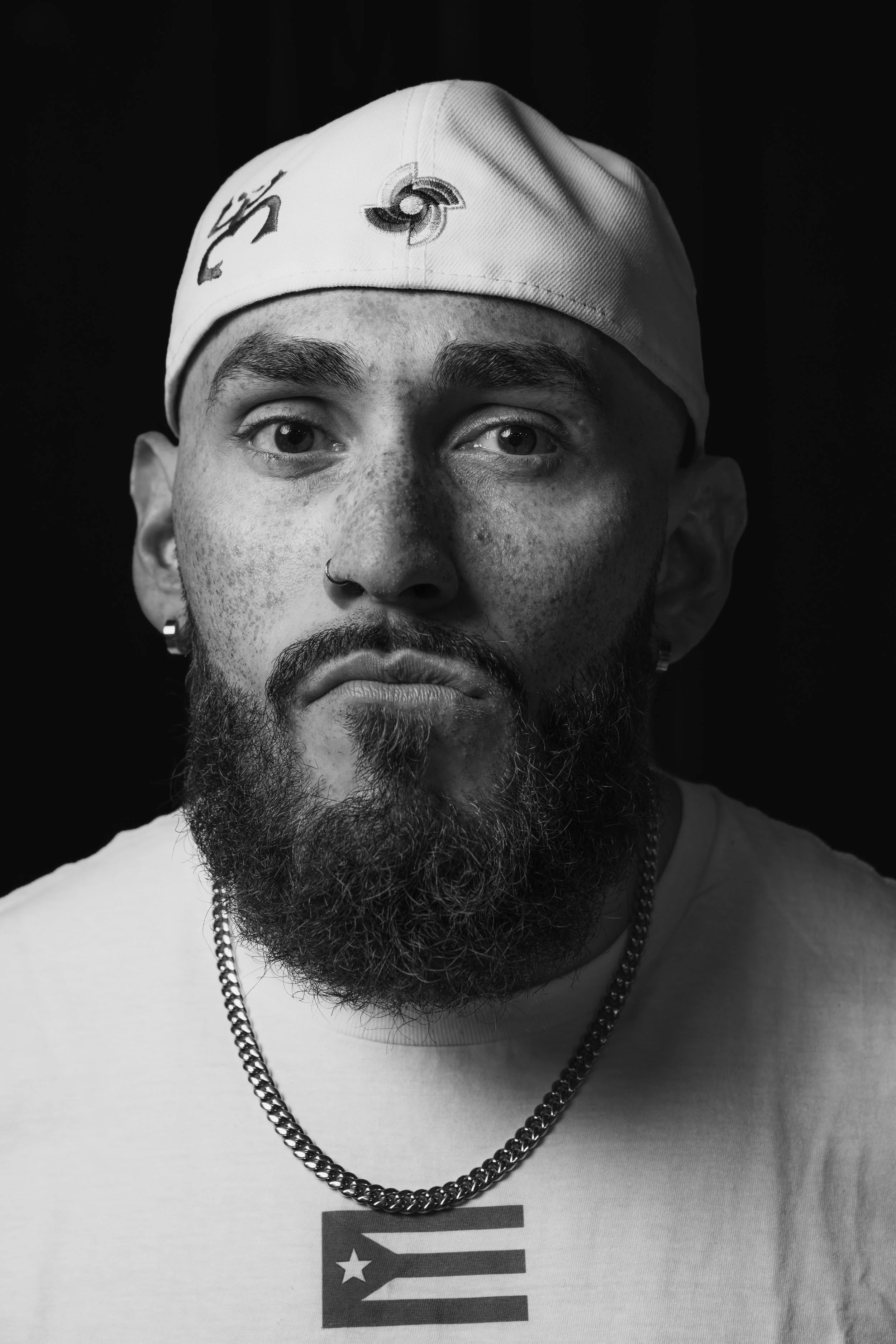 Close-up black and white photo of a man's face, wearing a bandana with logos, a chain necklace, and a T-shirt with a graphic of the American flag.