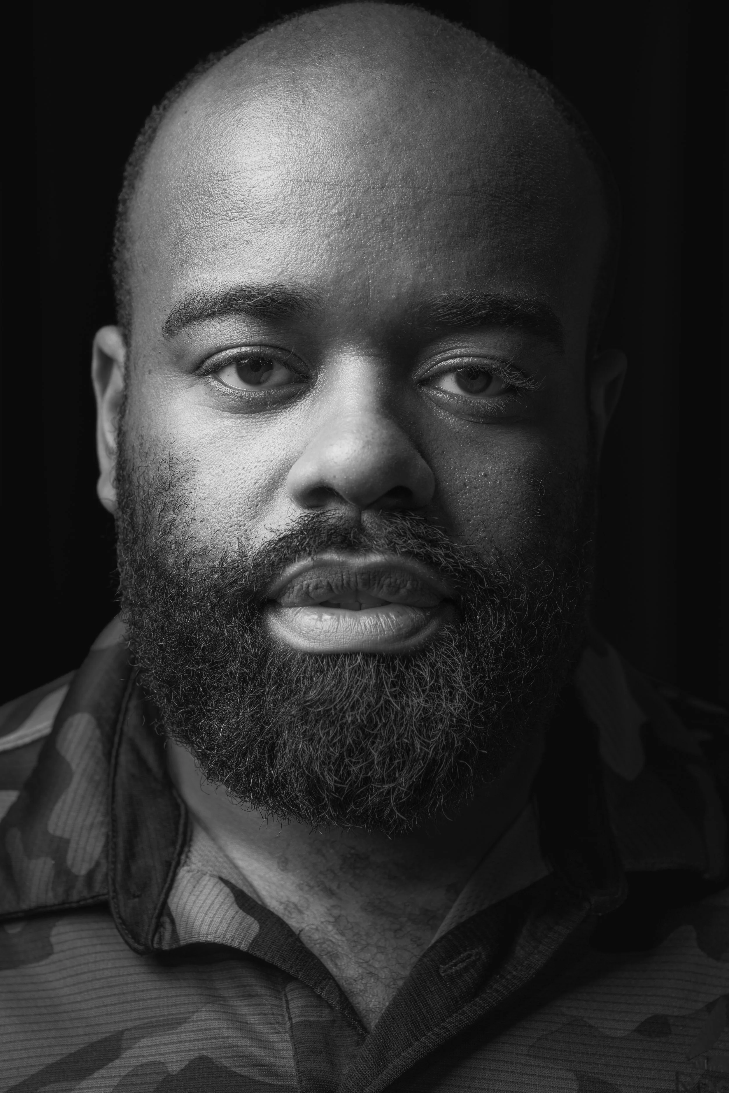 Close-up black-and-white portrait of a bearded man with short hair, wearing a striped shirt and a backpack strap visible on his shoulder.