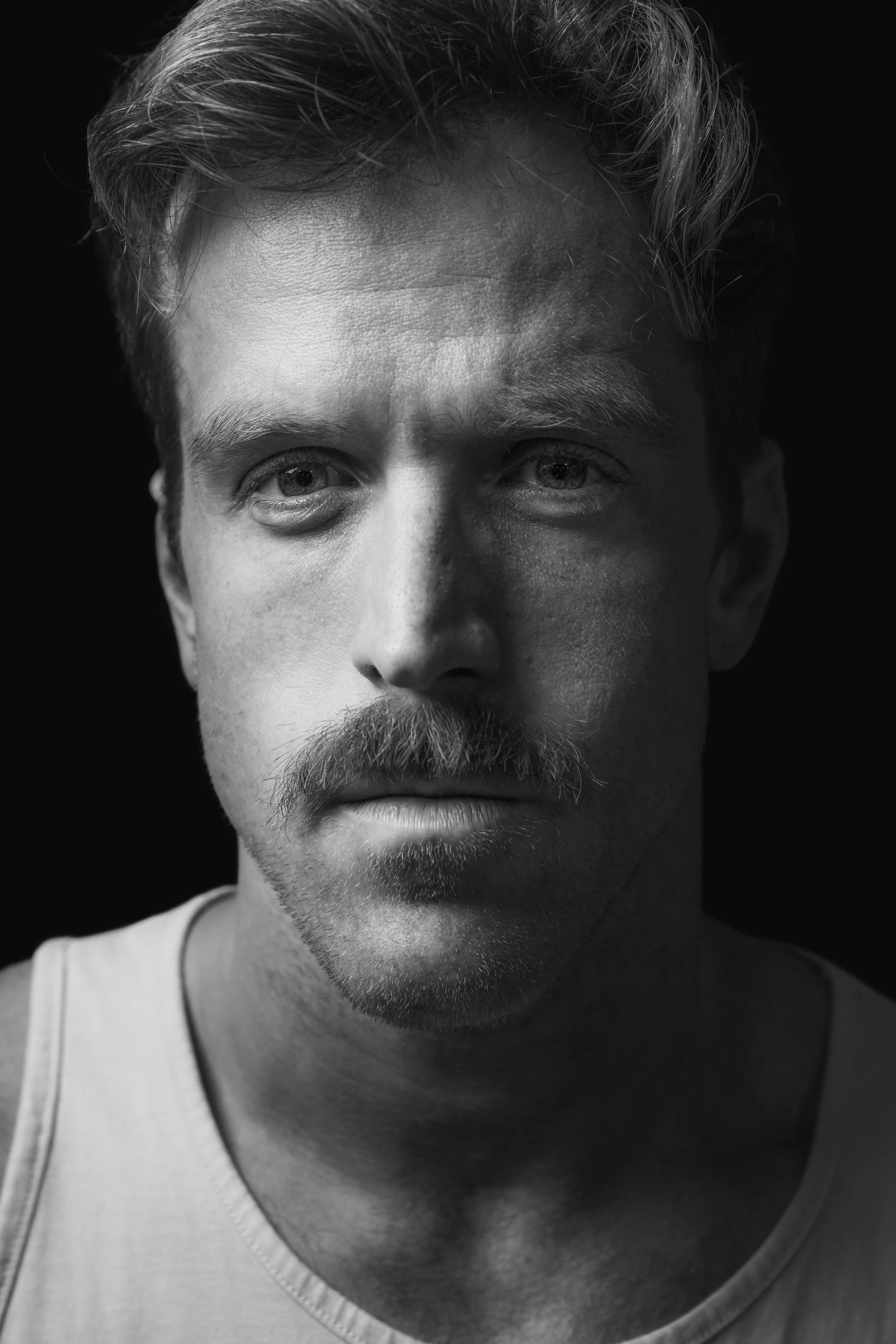 Close-up black and white portrait of a man with short, wavy hair, light facial hair, and wearing a tank top, against a black background.