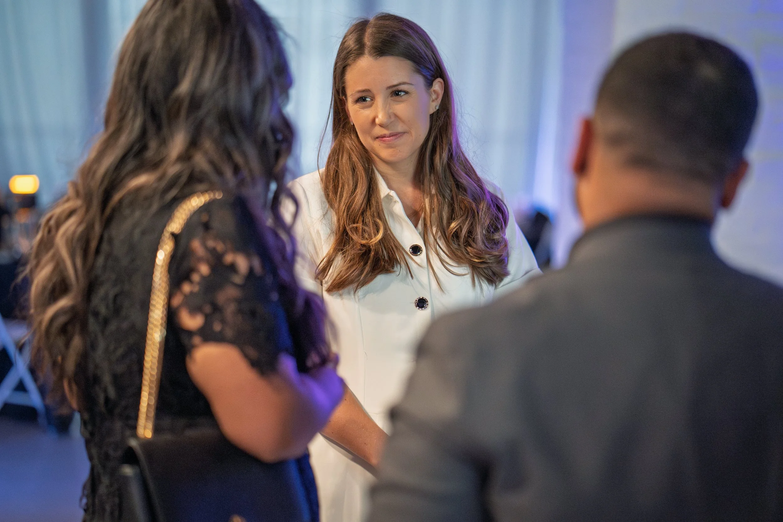 Three people in conversation at an indoor event, with the woman in the middle facing the camera. The woman in the foreground has long, wavy hair and wears a black lace top. The person on the right has short hair and is wearing a suit, and the person 