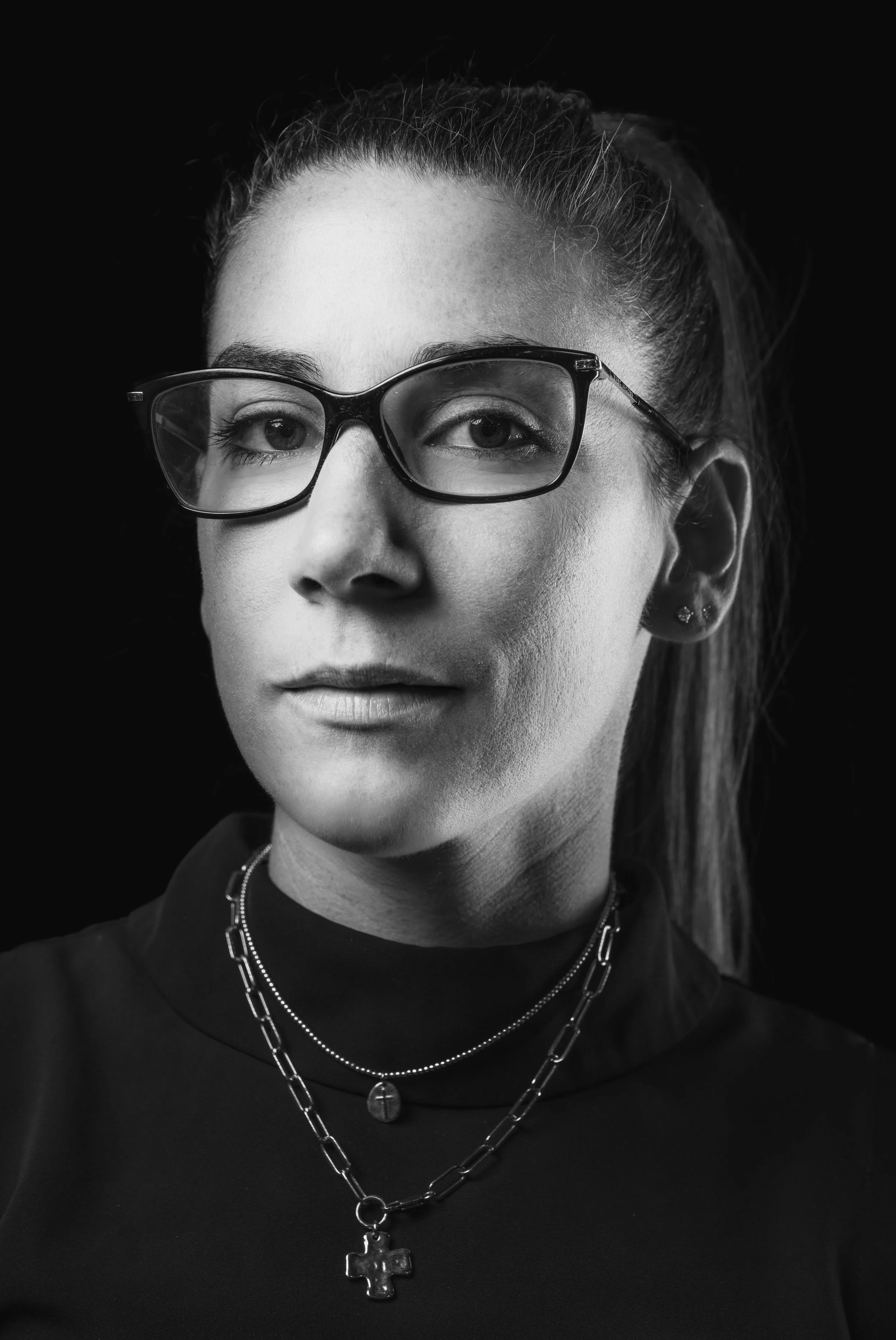 Black and white close-up portrait of a young woman wearing glasses, layered necklaces, and earrings, with her hair pulled back. She has light skin and a neutral facial expression.