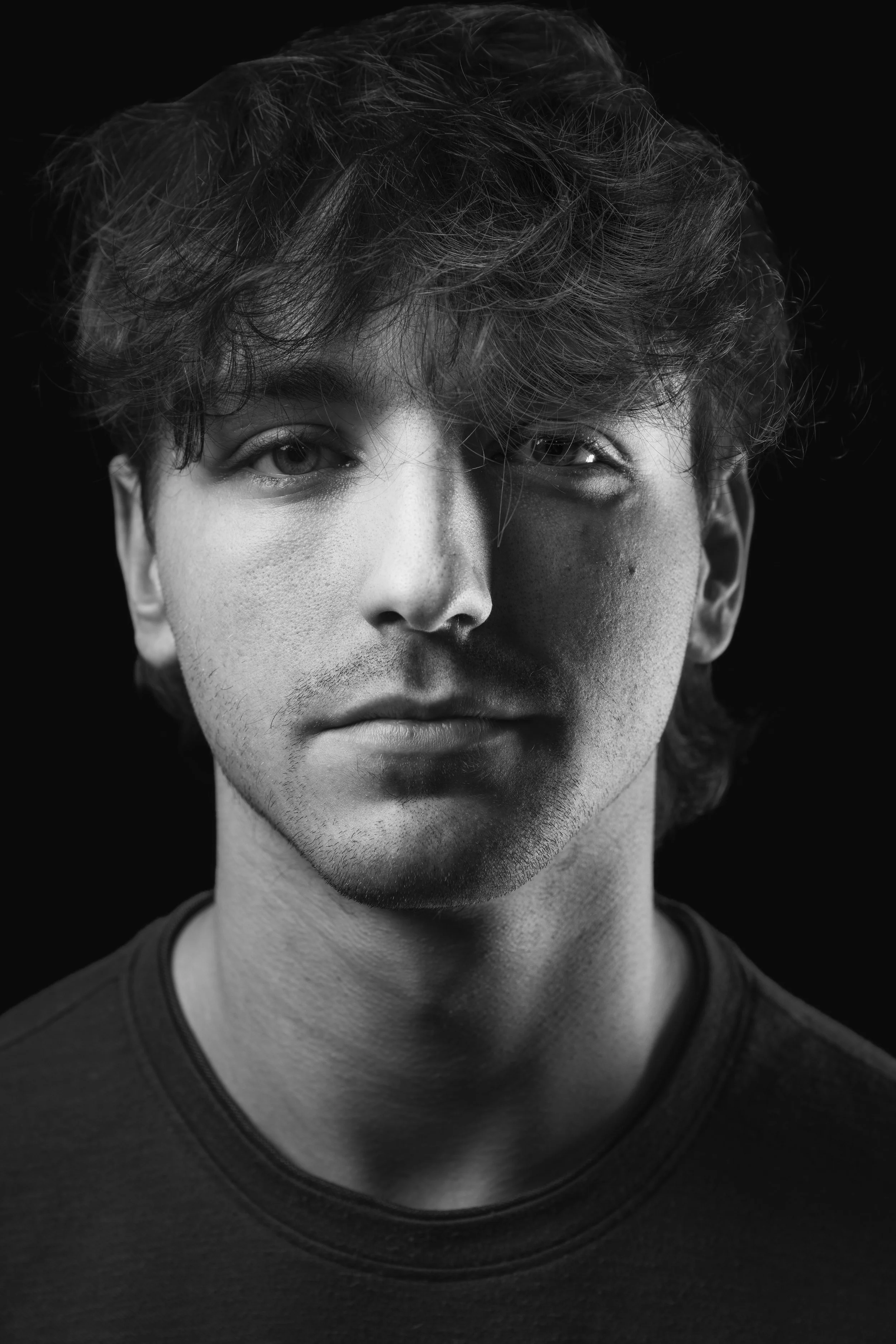 Close-up black and white portrait of a young man with curly hair, wearing a dark t-shirt, against a black background.