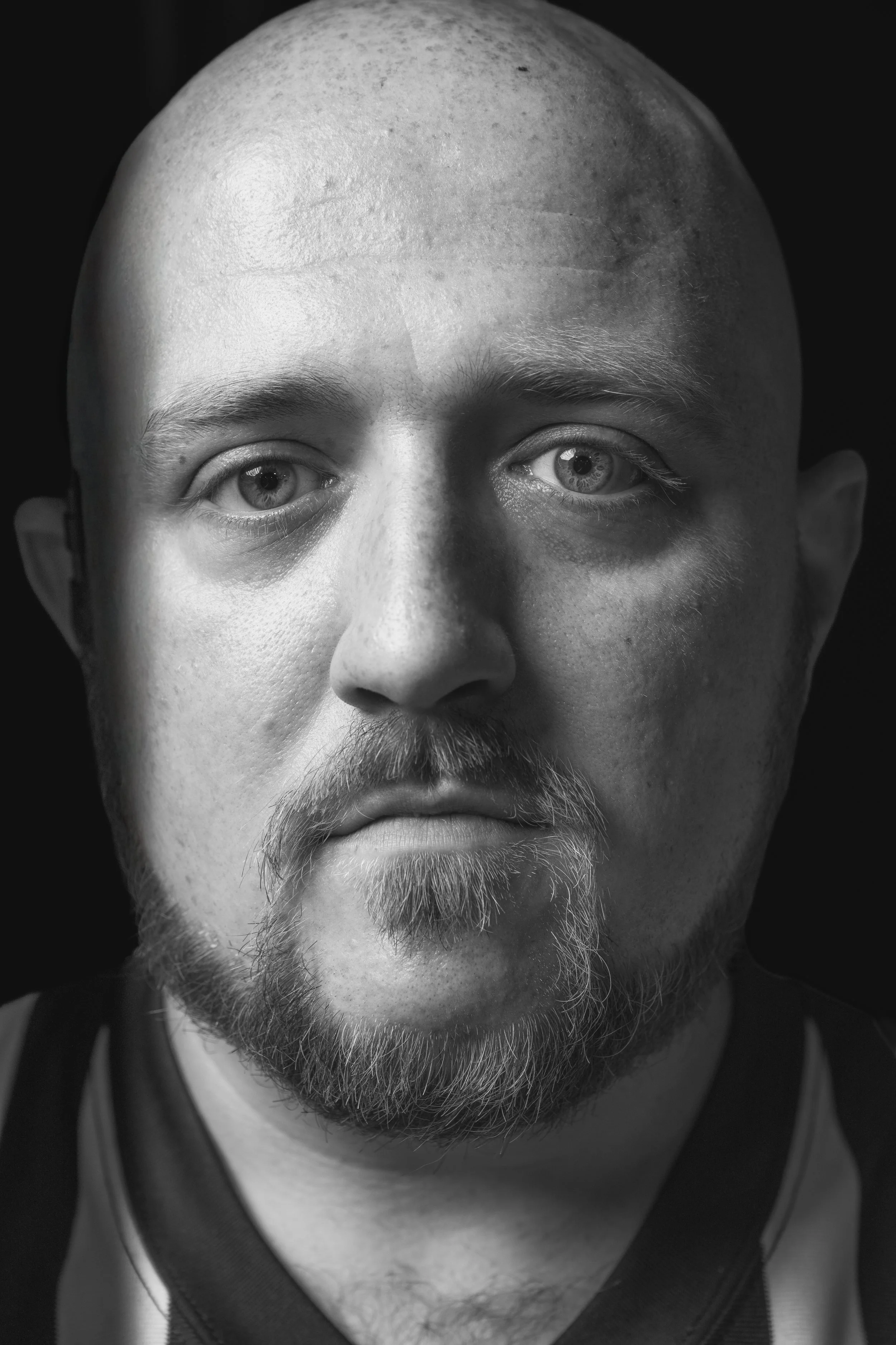 Close-up portrait of a man with a shaved head, light-colored eyes, facial hair, and wearing a sports jersey, against a dark background.