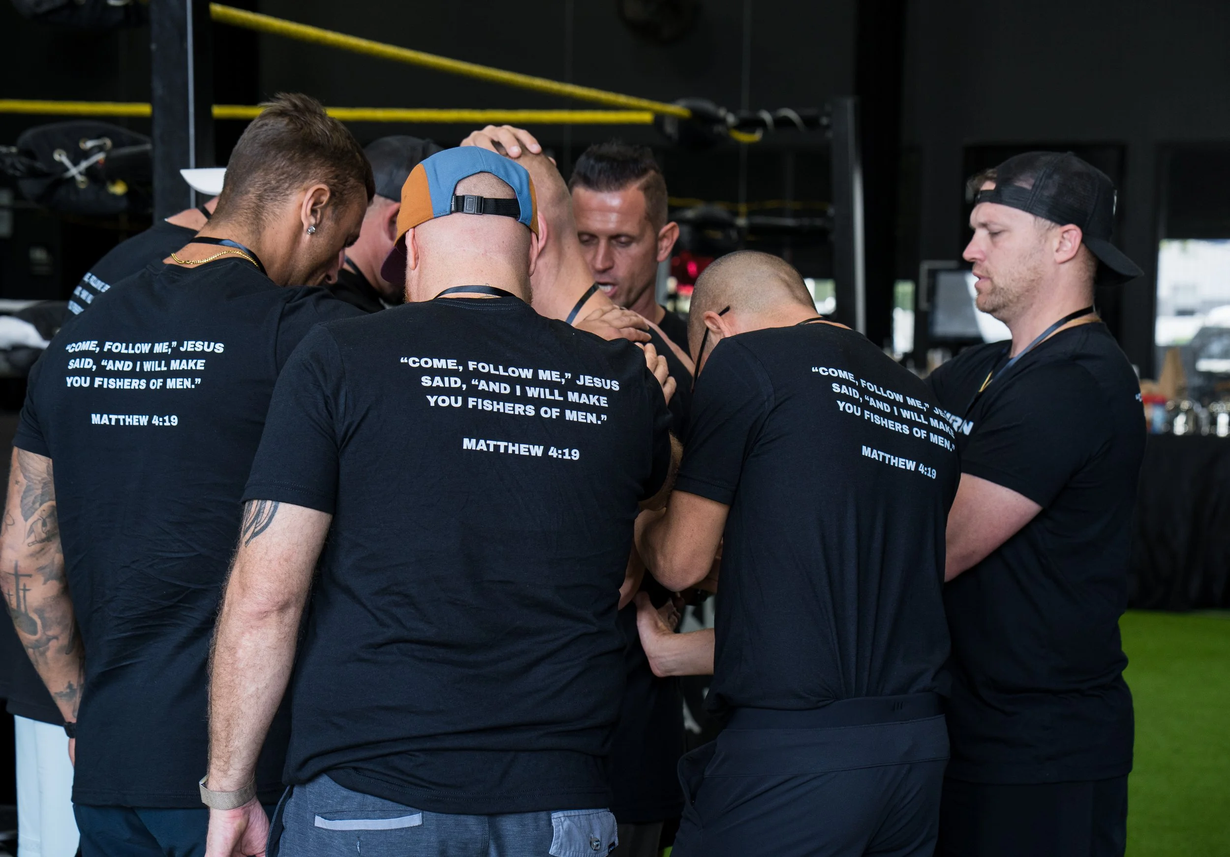 A group of men in black T-shirts gathered in a circle, praying together.
