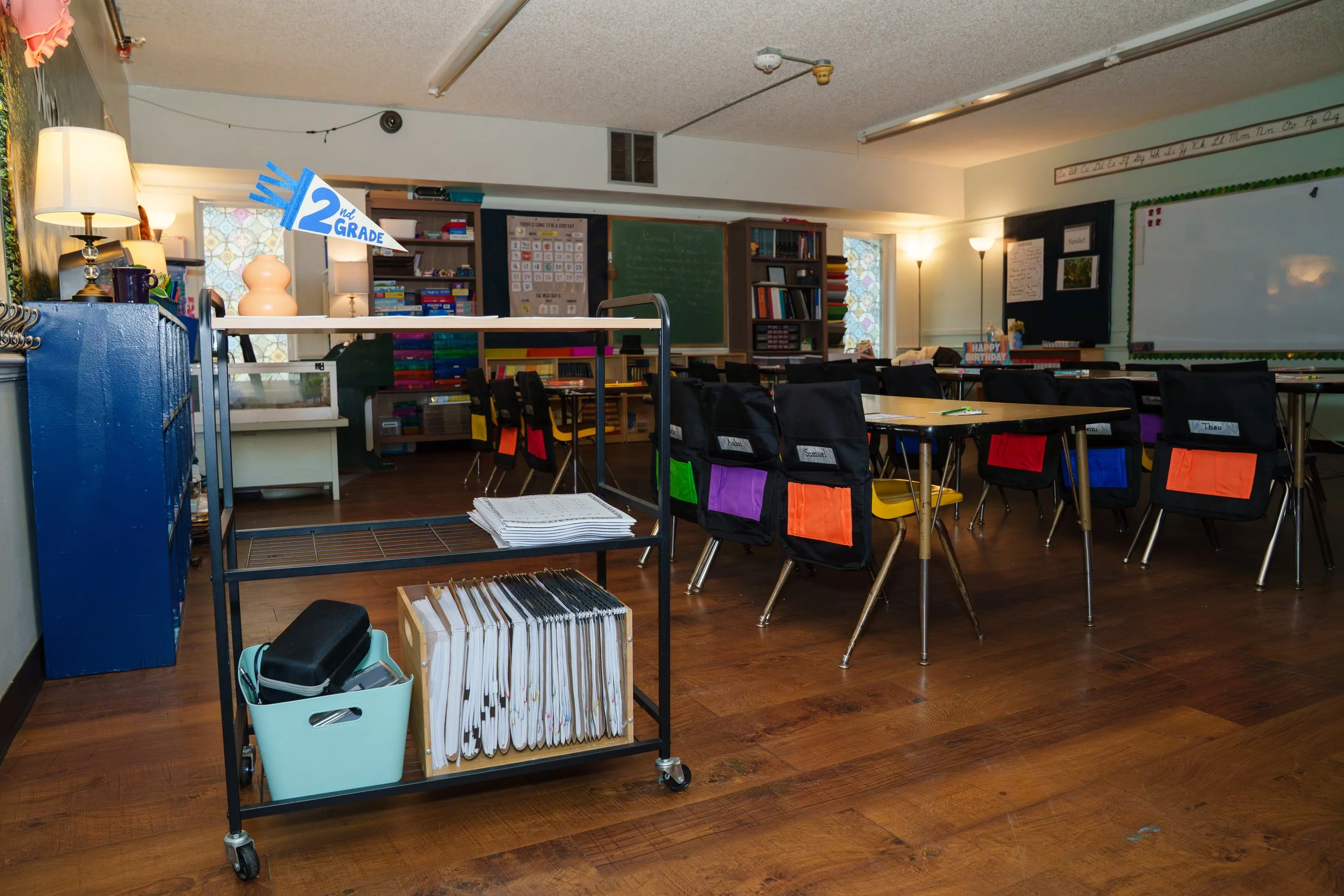 A classroom with a decorated bulletin board, a chalkboard, and desks with chairs. There are storage shelves filled with books and supplies, and a table with a sign that reads '2nd Grade.' Various items such as binders, a backpack, and a lamp are visi