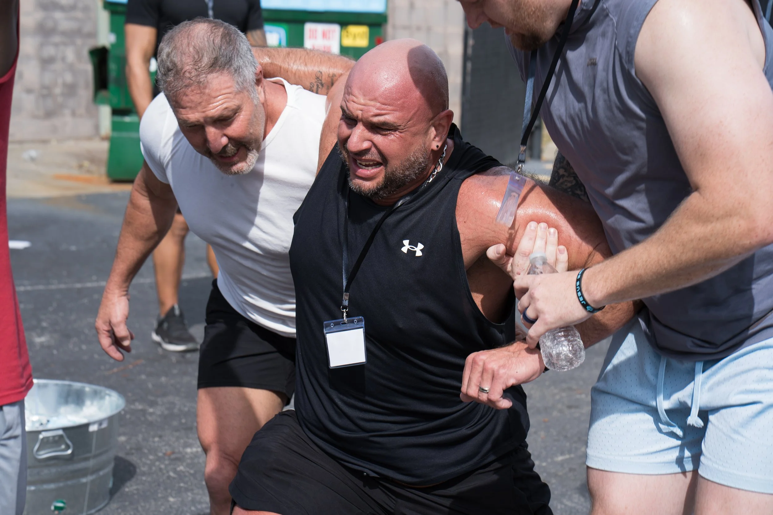 Three men assisting a man who appears to be experiencing a medical emergency or exhaustion, with one holding a water bottle and others supporting him, outdoors.