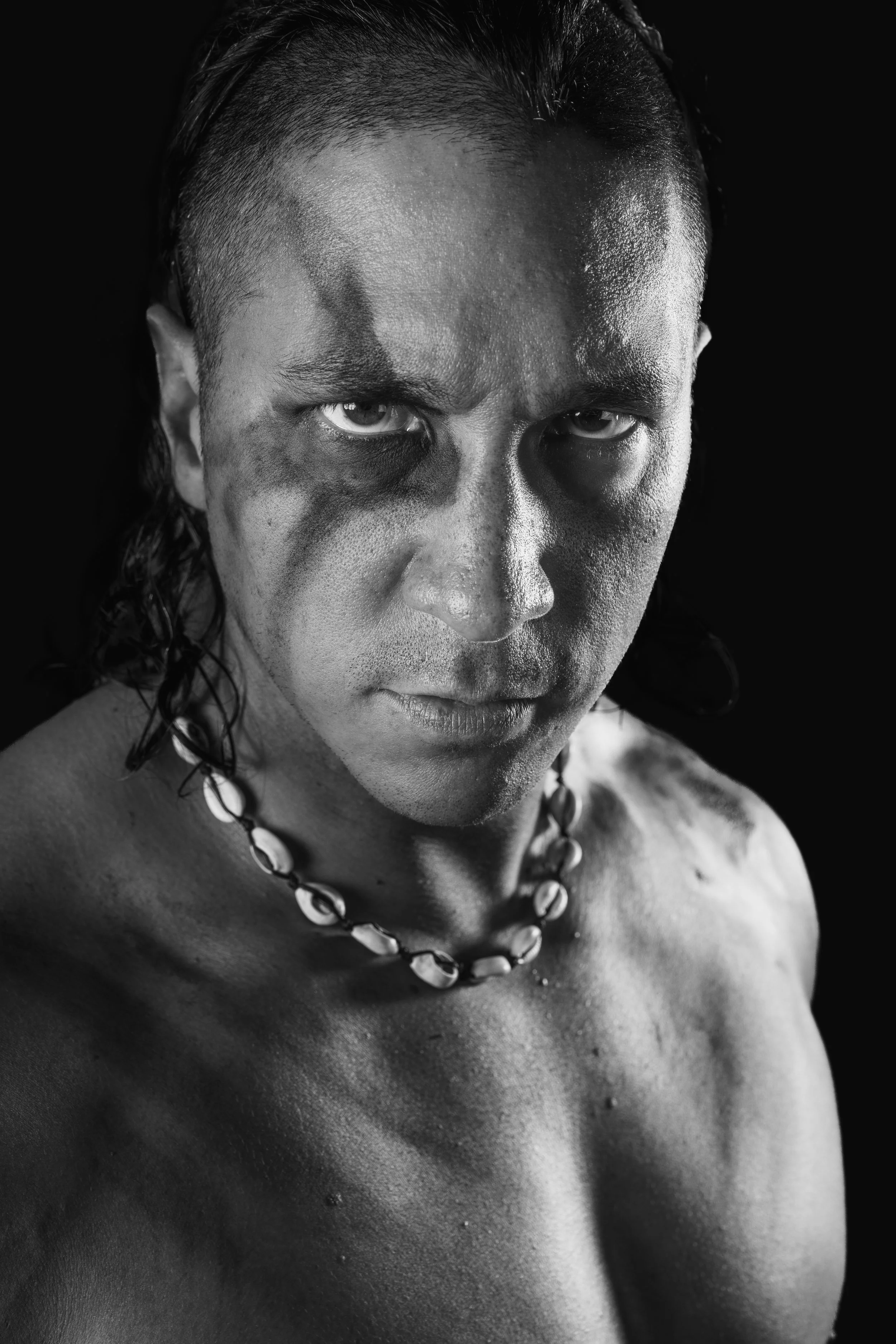 Close-up black and white photograph of a person's face and upper body, with water droplets visible on the skin, wearing a necklace made of shells or beads.