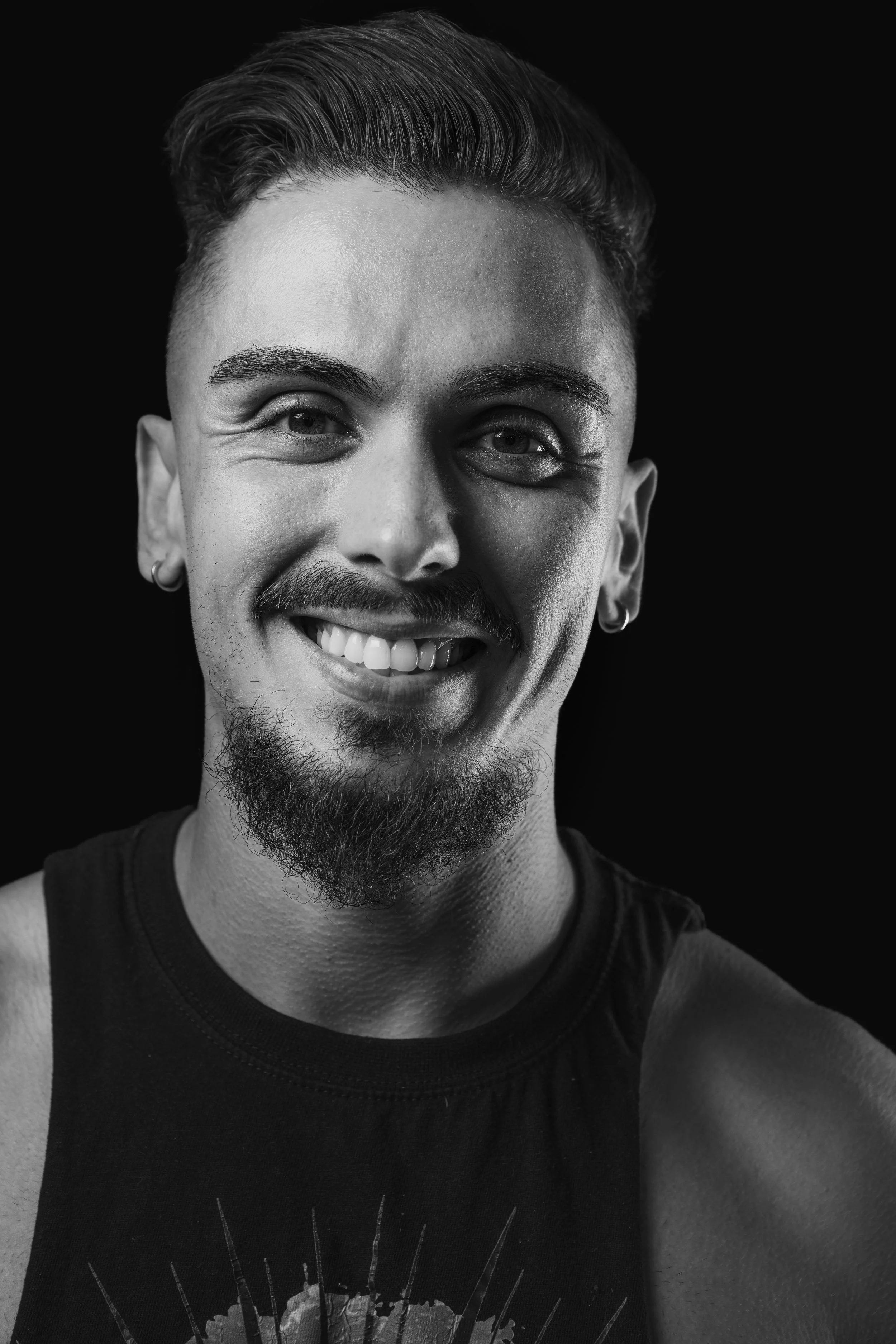 A black and white close-up portrait of a smiling man with short, styled hair, earrings, a mustache, and a goatee, wearing a sleeveless shirt with a graphic design.