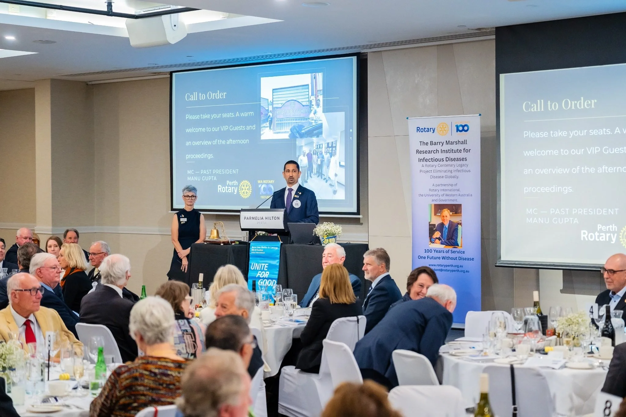 A formal event with a large attendance of mostly older adults, seated at round tables with white tablecloths, glasses, and bottles. A man in a suit stands behind a lectern with a sign that reads "Parmelia Hilton," speaking to the audience. Large screens display a presentation slide with the title "Call to Order" and information about a Rotary event, featuring logos of Rotary International and the 100 Years of Service. A woman stands near the speaker, and a vertical banner on the right side displays Rotary logos and details about the event.