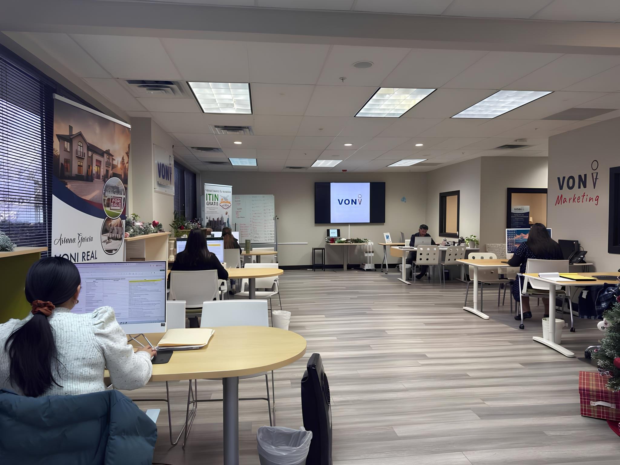 An office with multiple workers seated at desks, some working on computers, with a large screen displaying 'VON' in the background. There are promotional posters on the walls, holiday decorations, and windows with blinds.