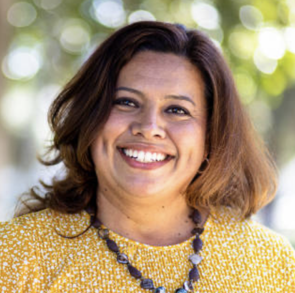 A woman with shoulder-length brown hair, smiling, outdoors with blurred green trees in the background, wearing a yellow patterned top and a beaded necklace.