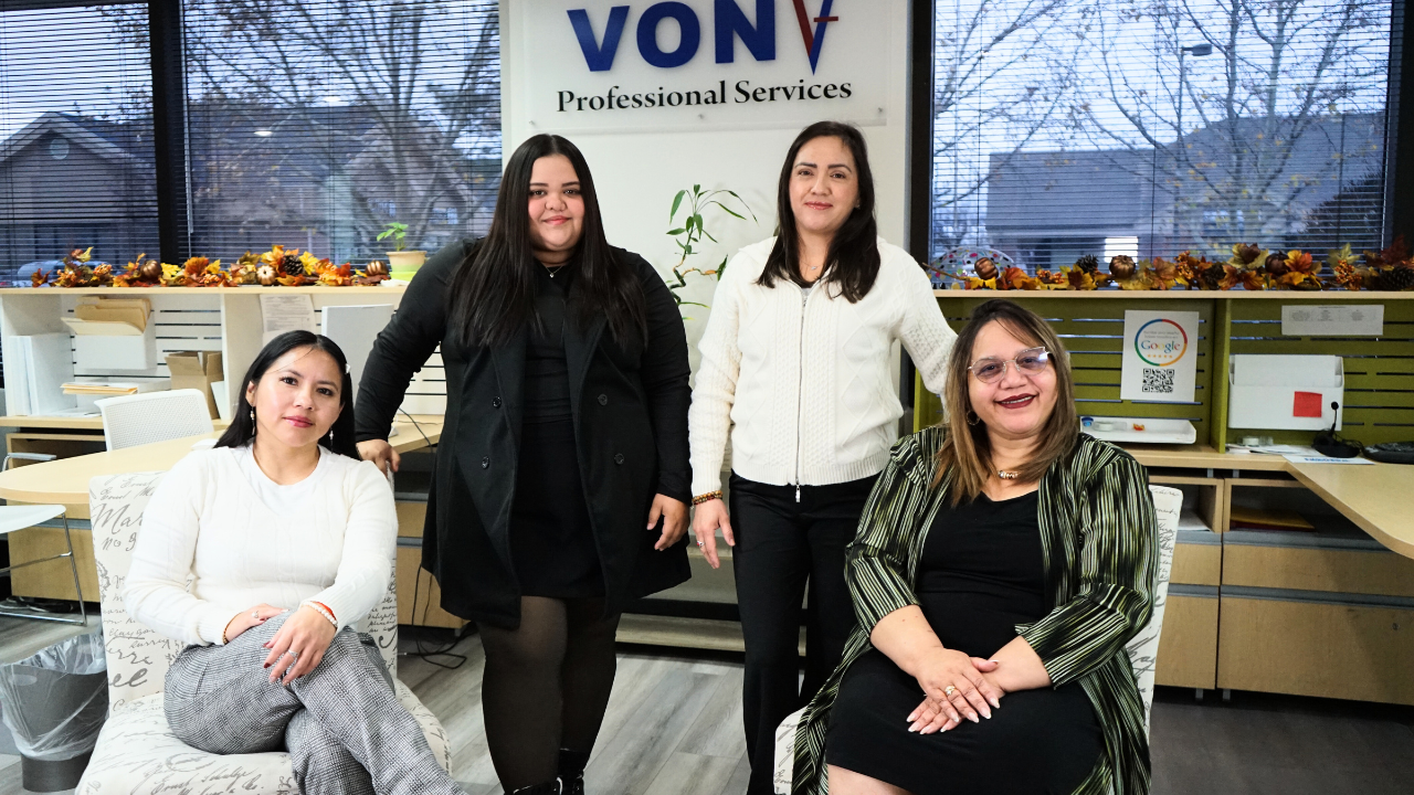 Four women in an office pose for a photo, two sitting and two standing, with a sign reading 'VONV Professional Services' in the background and windows showing trees and buildings outside.