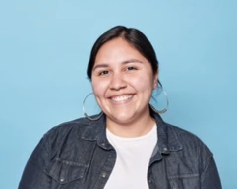 A woman smiling, wearing hoop earrings and a denim jacket against a blue background.