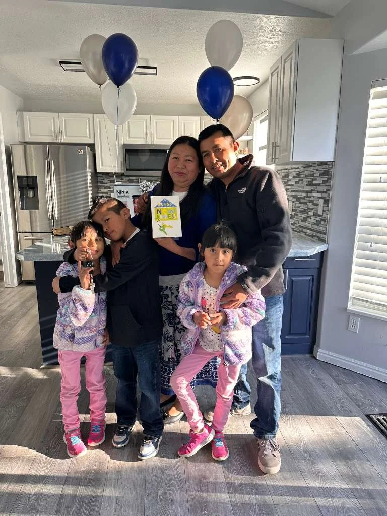 A family of five standing inside a modern kitchen with white cabinets and stainless steel appliances, holding balloons and a greeting card, with sunlight coming through a window.