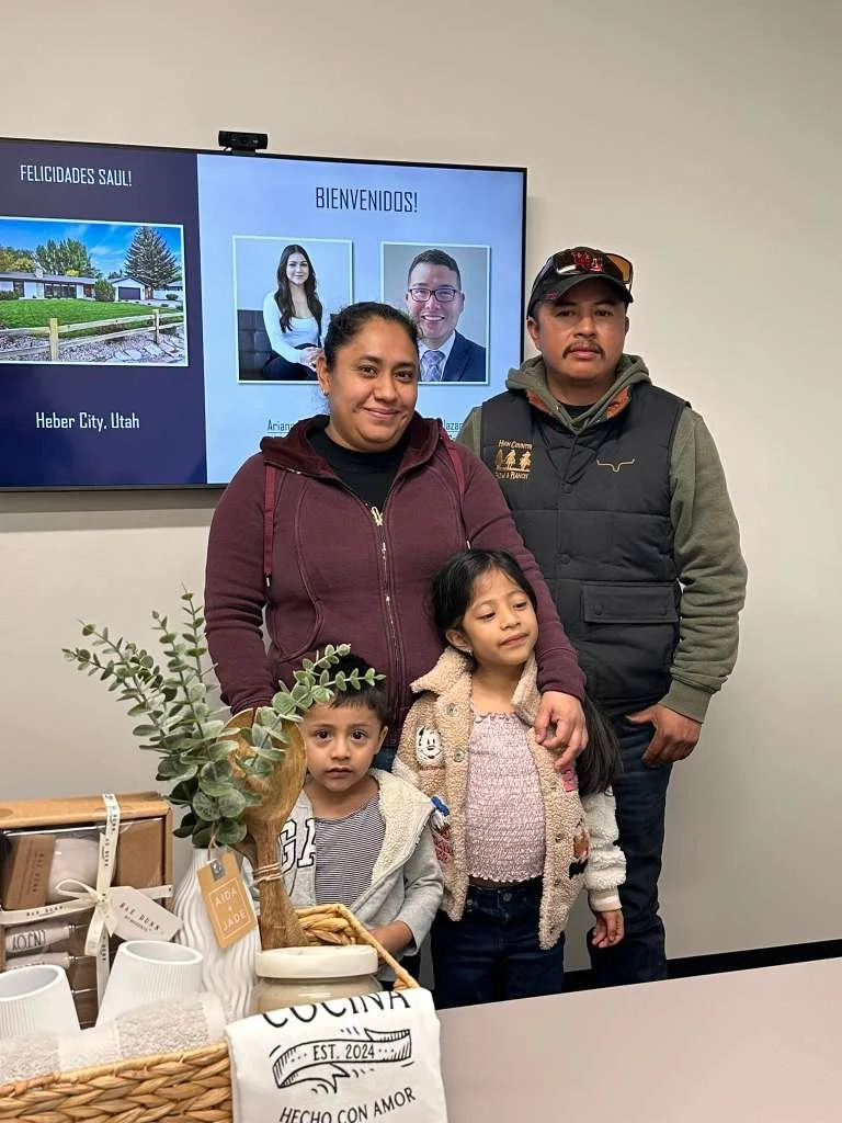 Family of four, including two children and two adults, posing indoors in front of a digital display with welcome messages and portraits. There are gift baskets and decorative items on the table in front of them.