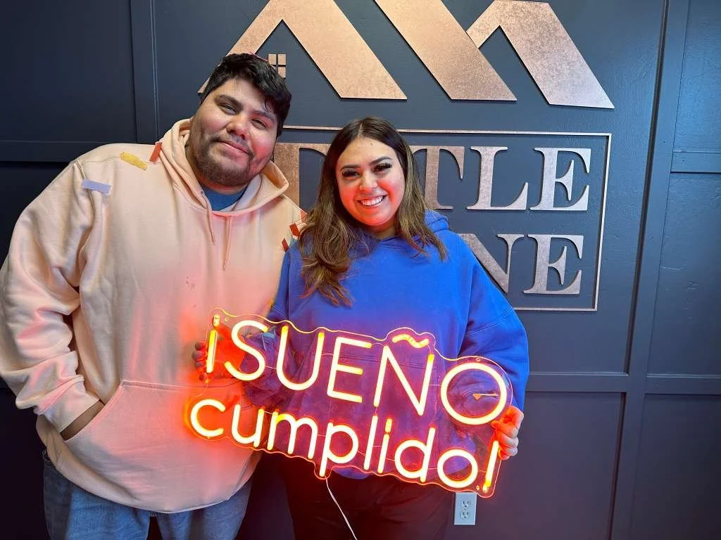 A smiling man and woman standing in front of a wall with a logo that reads 'DUSTLE' and holding a neon sign that says '¡SUENO cumplido!' which means 'Dream Come True' in Spanish.