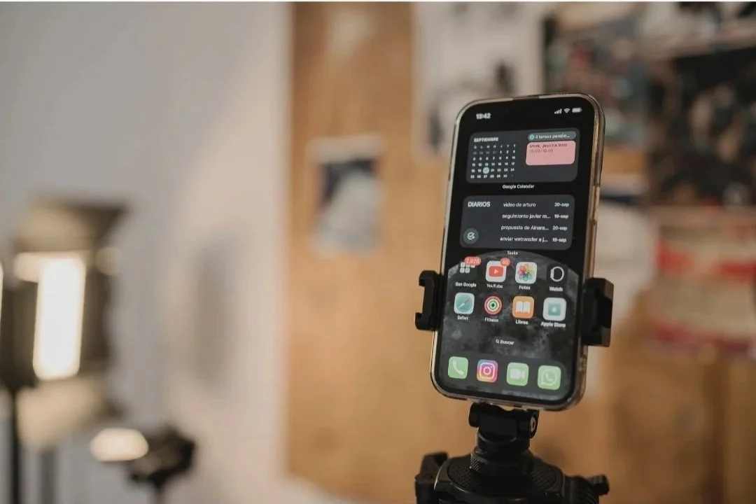 Smartphone mounted on a stand displaying home screen apps and notifications in a room with shelves.