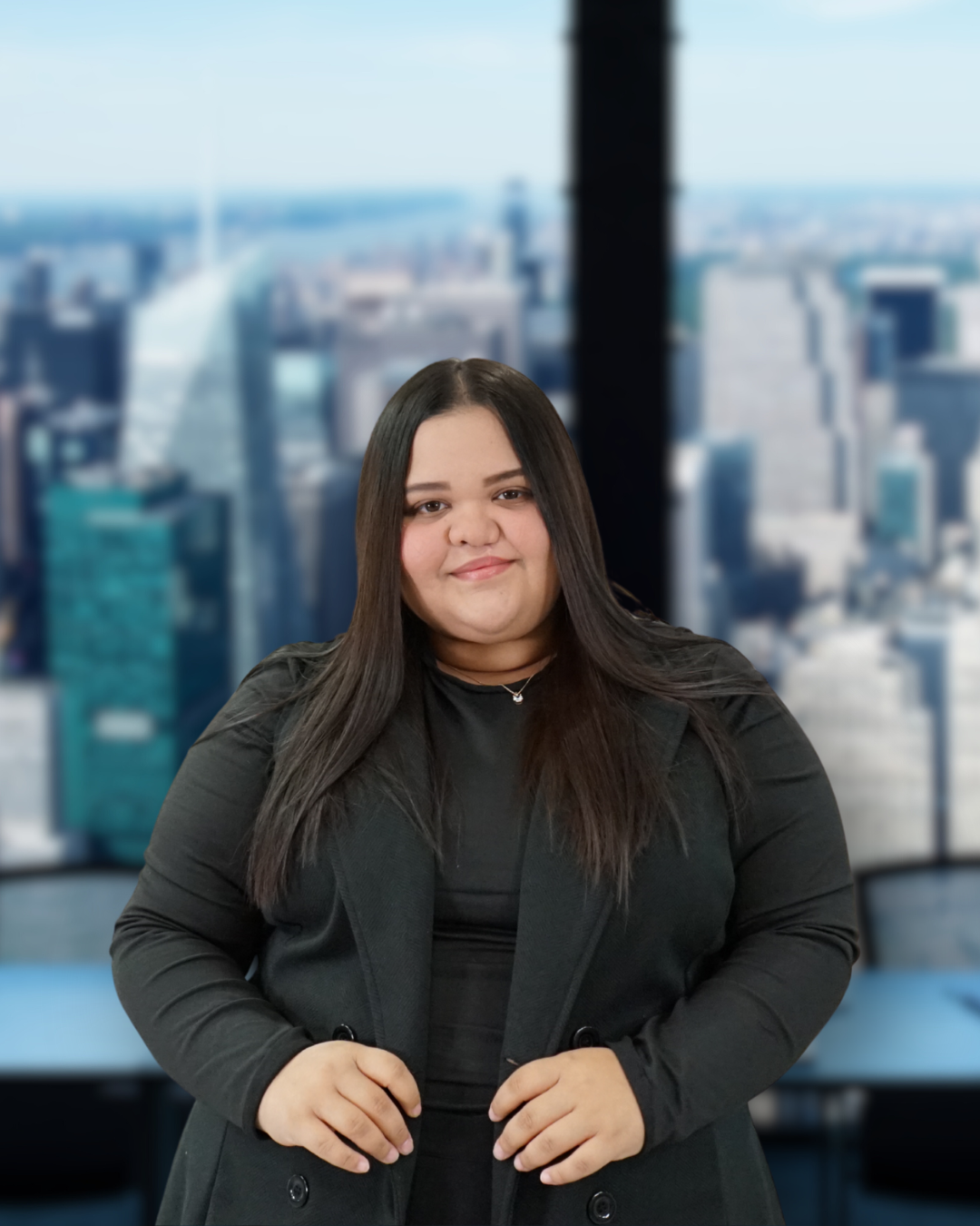 A woman with long dark hair wearing a black blazer and black top, standing in front of a large window overlooking a cityscape with tall buildings.