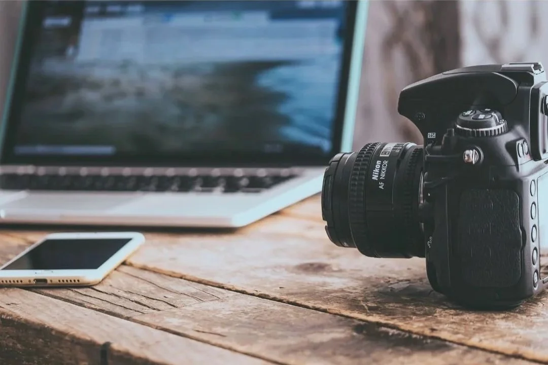A DSLR camera, a laptop, and a smartphone placed on a rustic wooden table.