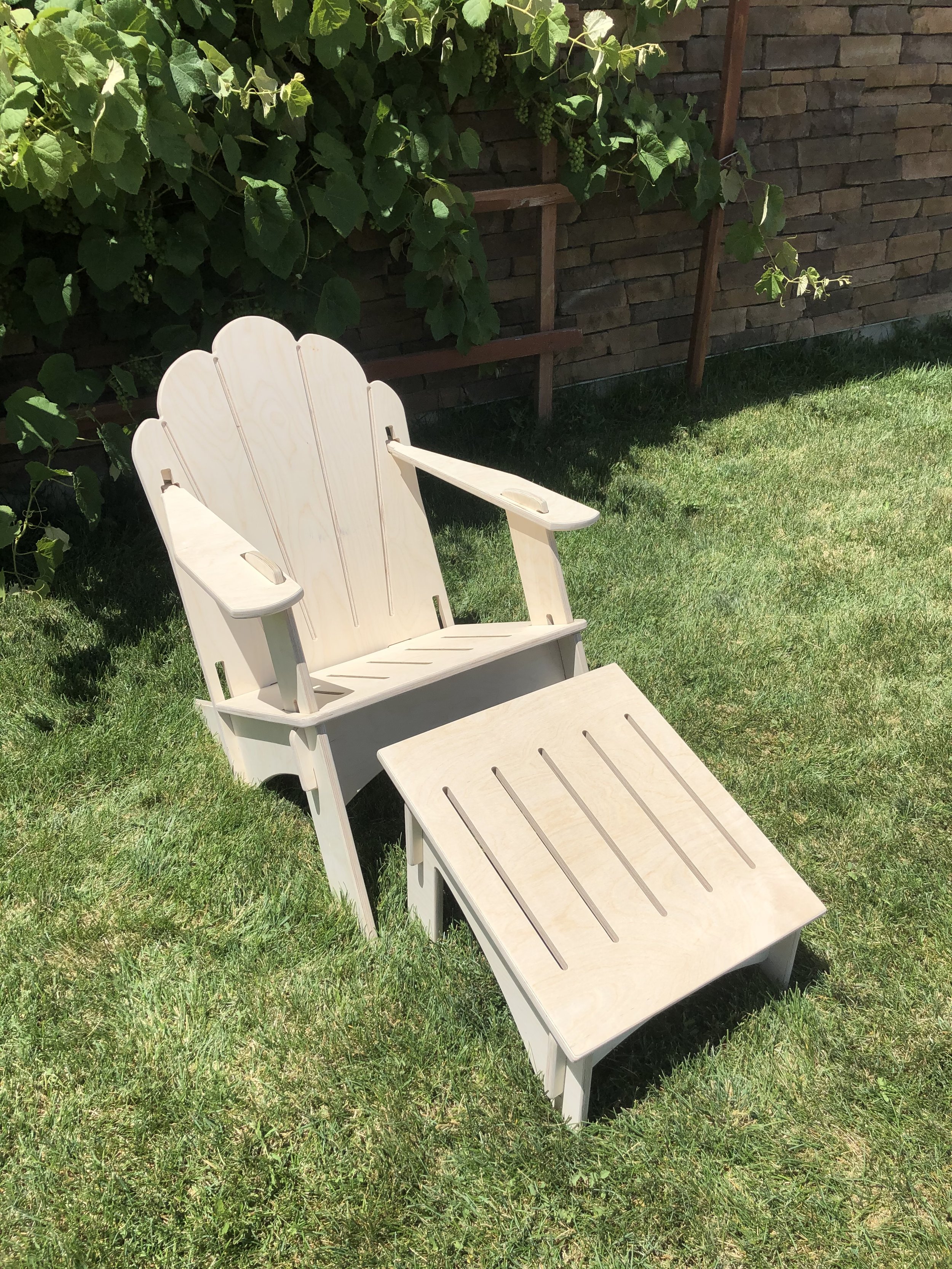 Natural wood Adirondack chair in a traditional style with a foot stool situated in green grass. Baltic Balsam wood finish to a comfy and collapsible chair