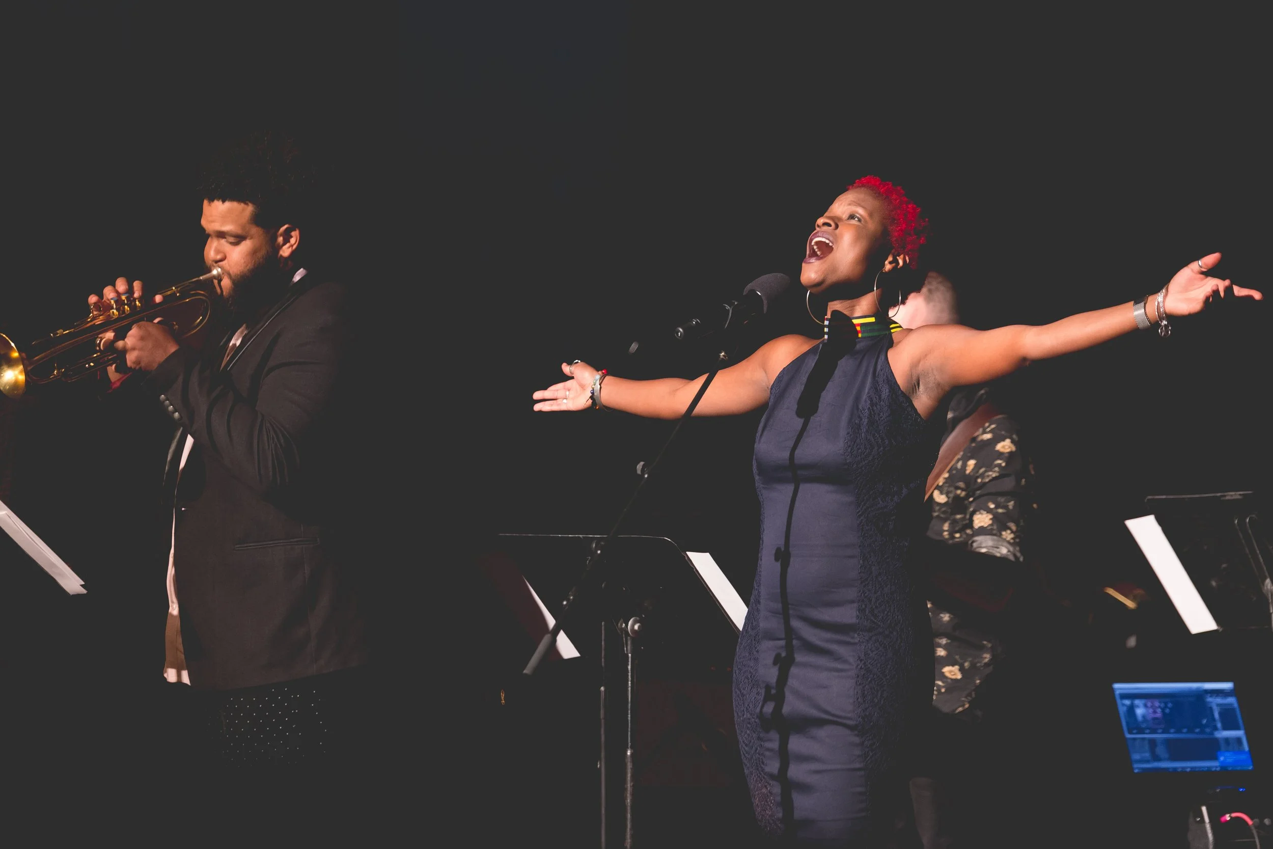 A female singer with red curly hair passionately singing with arms outstretched, standing in front of a microphone on a stage. To her left, a male jazz musician with curly hair plays a trumpet. A pianist is partially visible in the background. Darkne