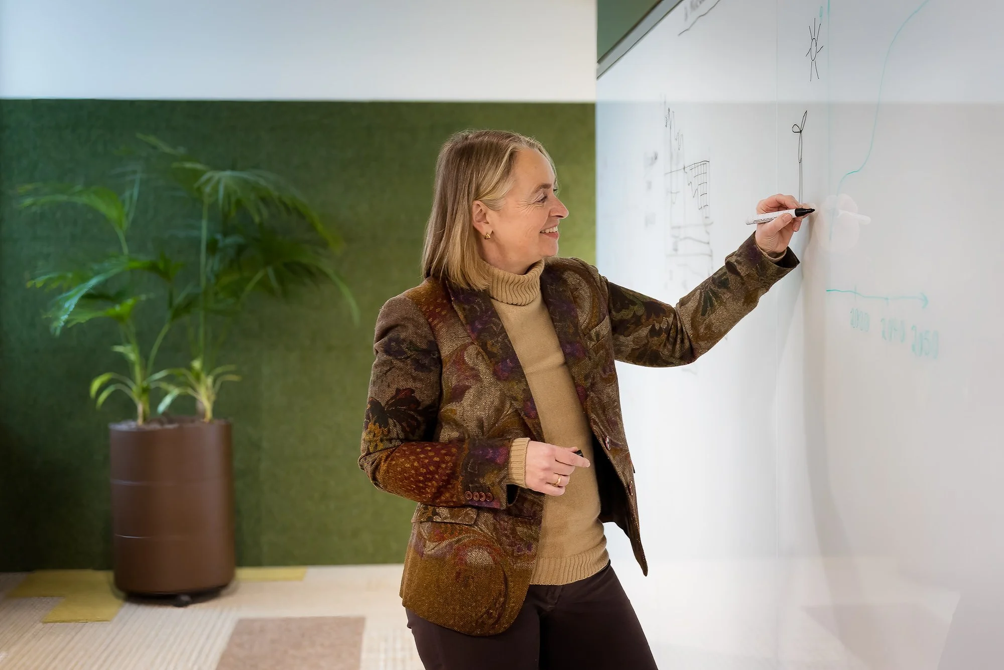 Eine Frau schreibt auf eine weiße Tafel in einem modernen Büro. Im Hintergrund ist eine grüne Wand und eine große Topfpflanze.