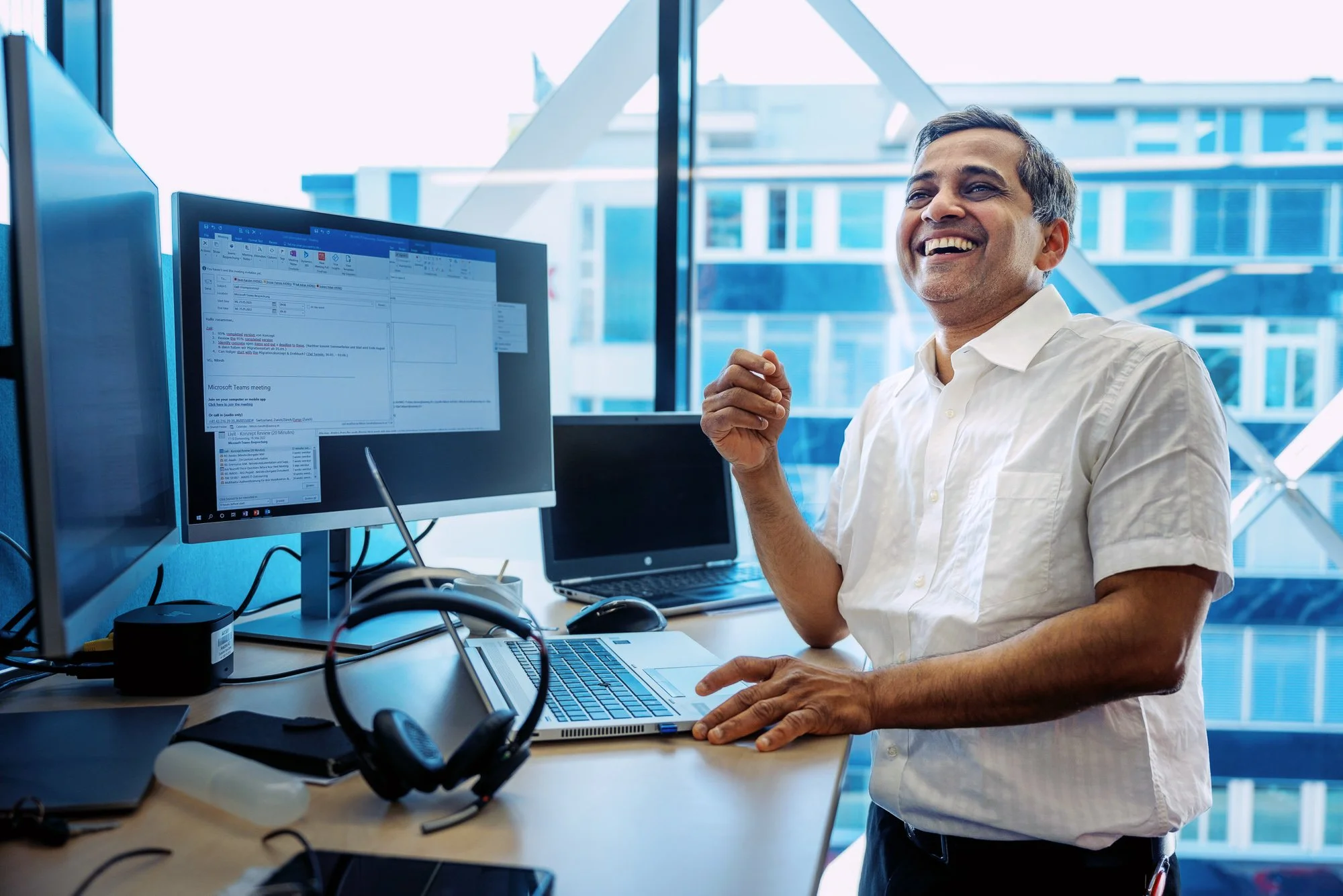 Ein Mann in einem weißen Hemd lächelt vor mehreren Computerbildschirmen in einem modernen Büro mit großen Fenstern.