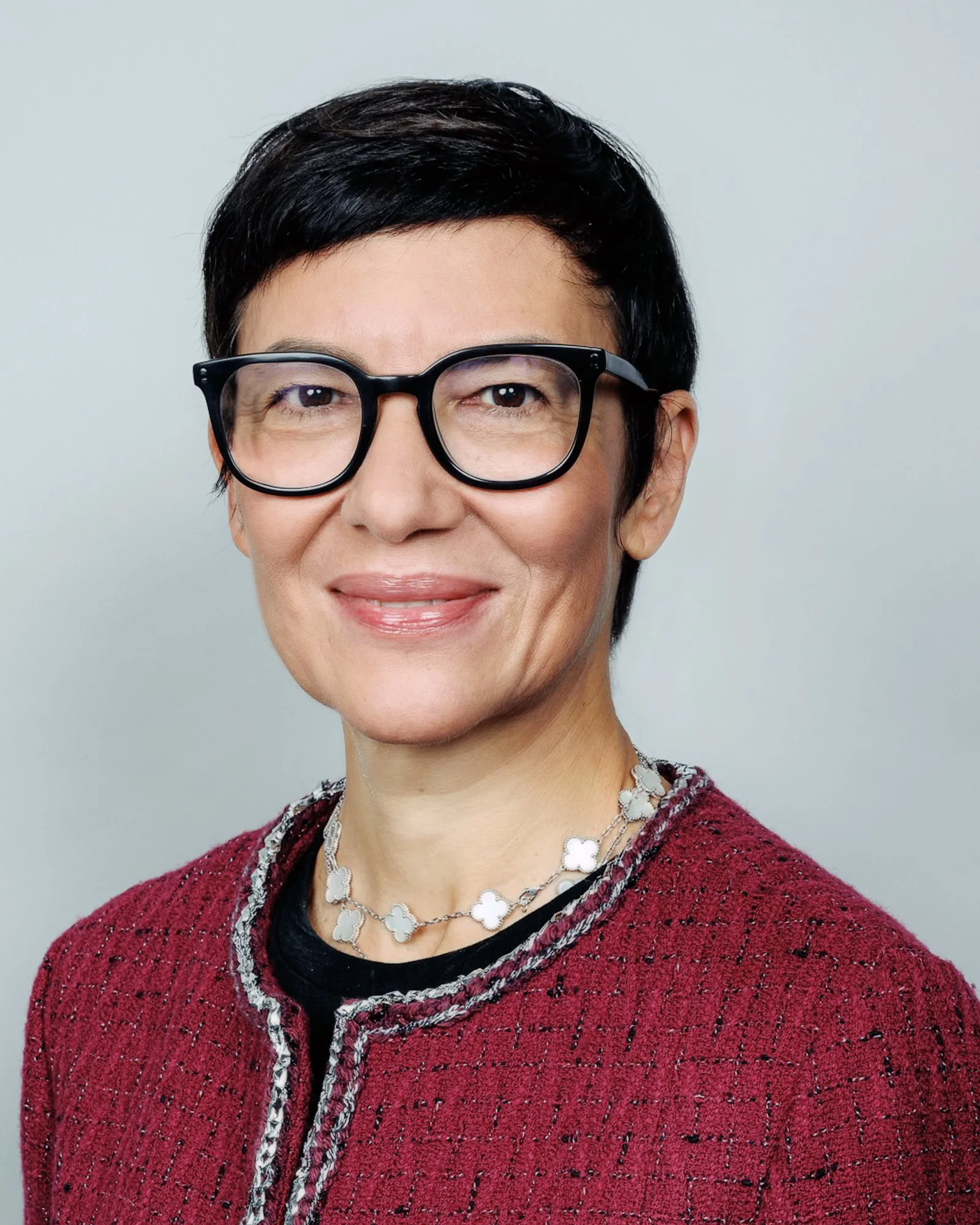 Porträt einer Frau mit kurzen schwarzen Haaren, schwarzen Brillen, rotem Jackett, schwarzem Oberteil und silberner Kette, vor neutralem Hintergrund.