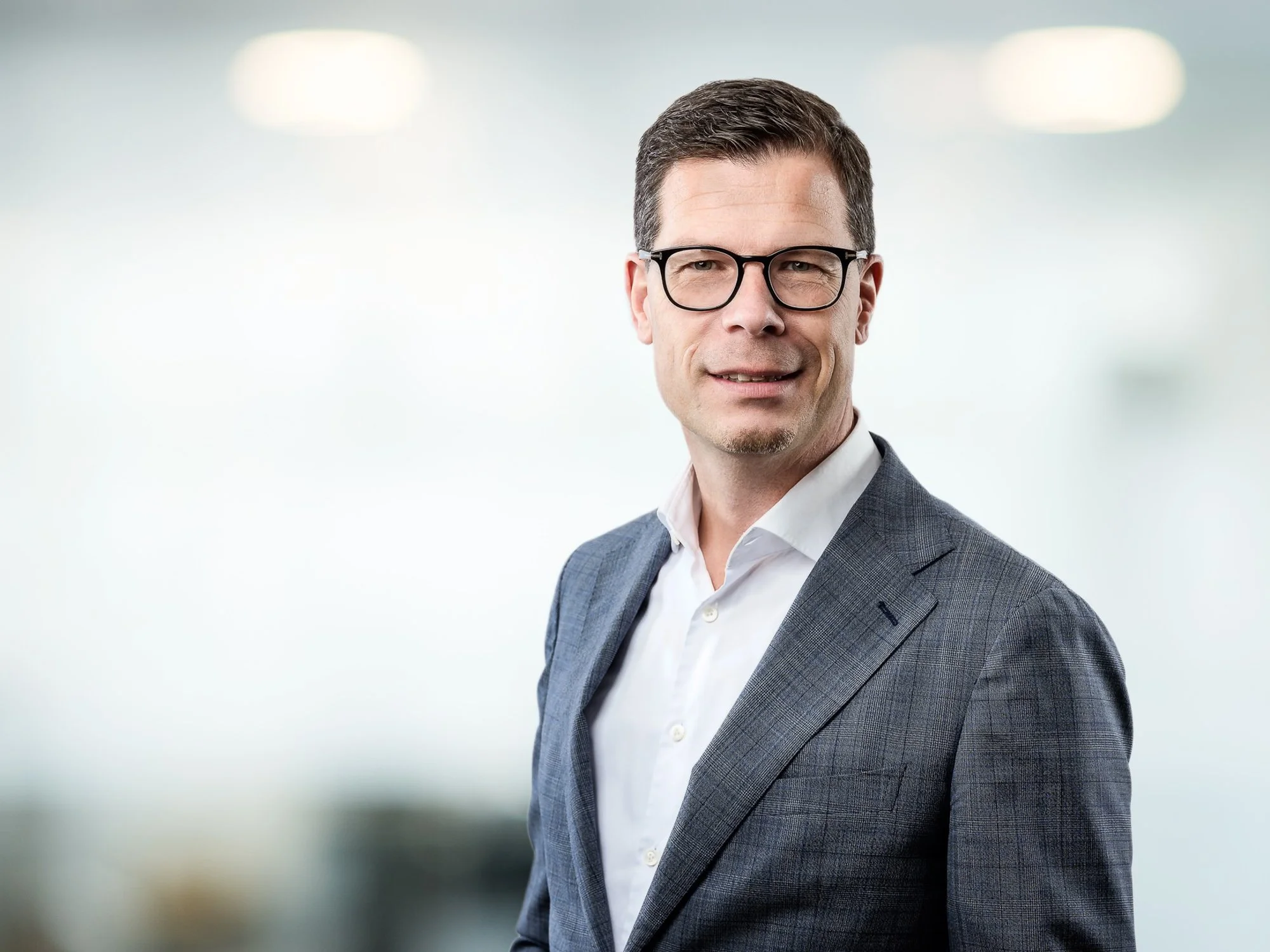 Geschäftsmann in Brille und Anzug vor einem hellen, unscharfen Hintergrund.