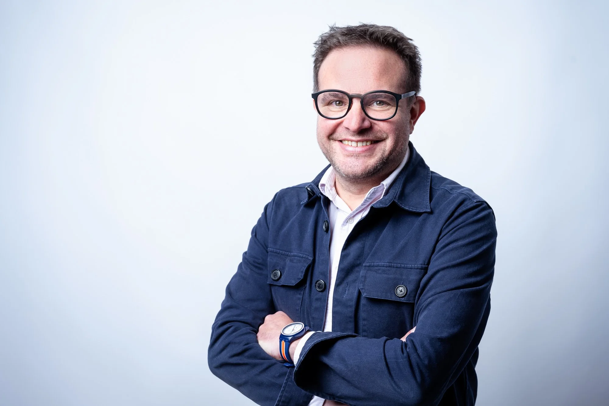 Ein lächelnder Mann mit Brille trägt eine dunkelblaue Jacke und eine Uhr, steht vor einem weißen Hintergrund.