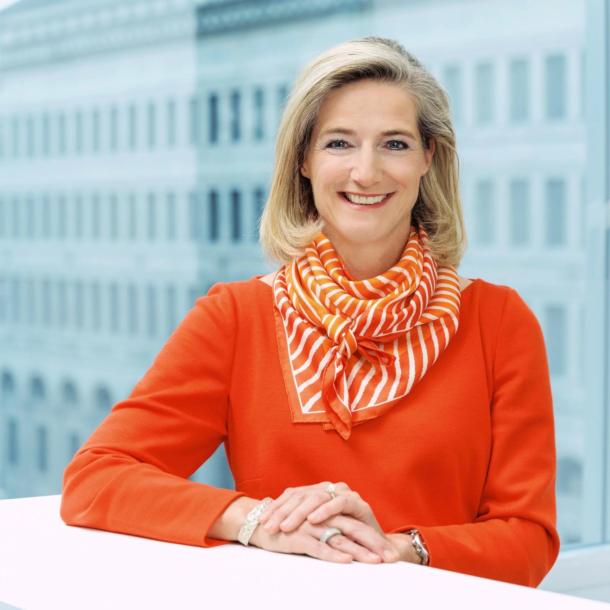 Eine lächelnde Frau mit blonden Haaren trägt ein orangefarbenes Kleid und ein orange-weiß gestreiftes Halstuch, sitzt an einem weißen Tisch vor einem Fenster mit Blick auf ein modernes Gebäude.