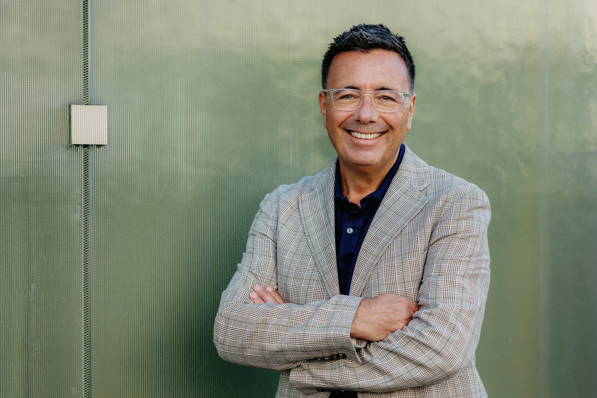 Ein lächelnder Mann in Brille trägt einen karierten Blazer und eine dunkle Hemdseite, steht mit verschränkten Armen vor einer grünen Metallwand.