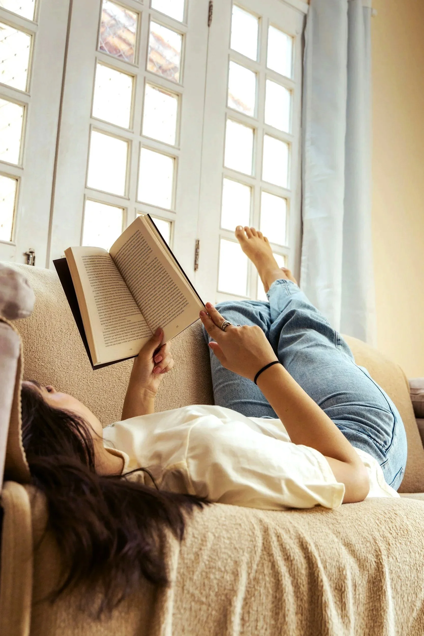 Person lying on a bed reading a book near a large window with white grid panes, with sunlight streaming in.