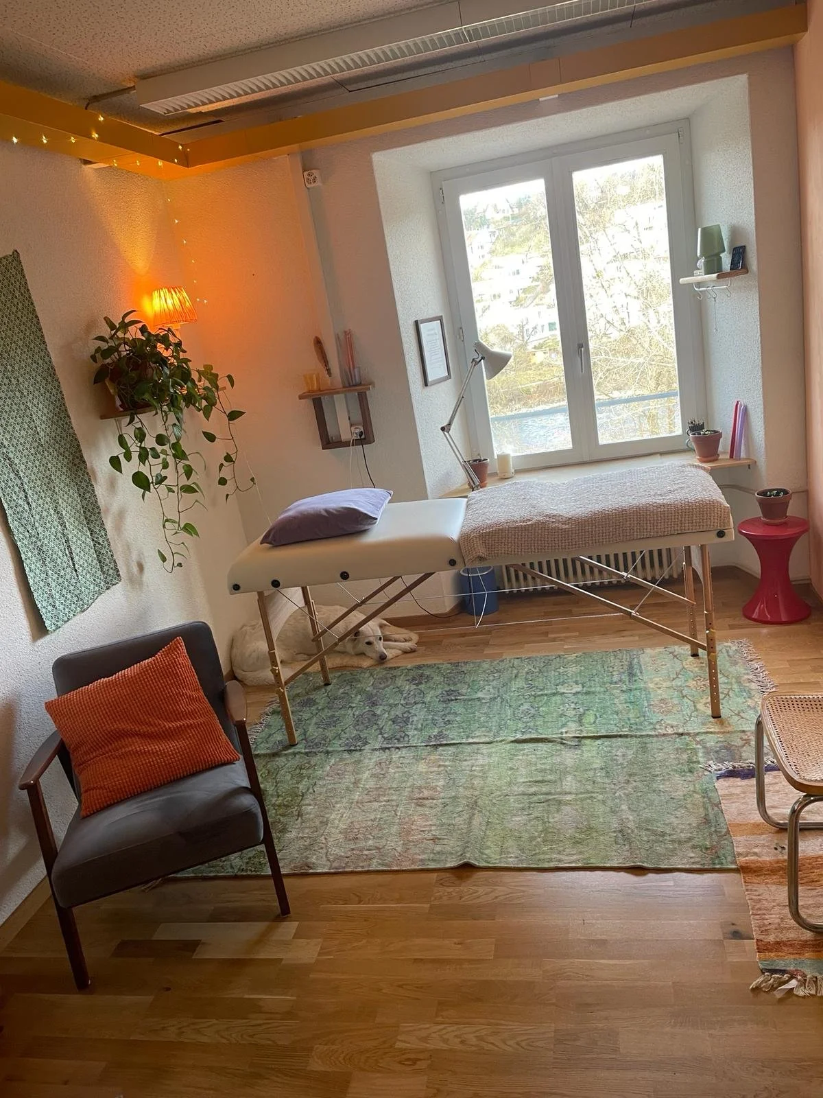 Ein Behandlungsraum mit Therapie- oder Massagetisch vor einem Fenster. Es gibt einen schwarzen Sessel mit orangener Kissen rechts, eine grüne Wanddeko, eine Pflanze, einen kleinen roten Tisch und mehrere Lichter und Dekorationen.