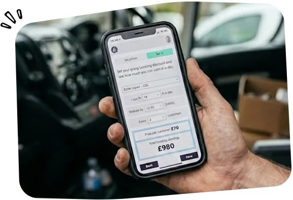 Hand holding a smartphone with a financial app open, displaying budget details and a total expense of 980 pounds on a vehicle in what appears to be a garage or vehicle service area.