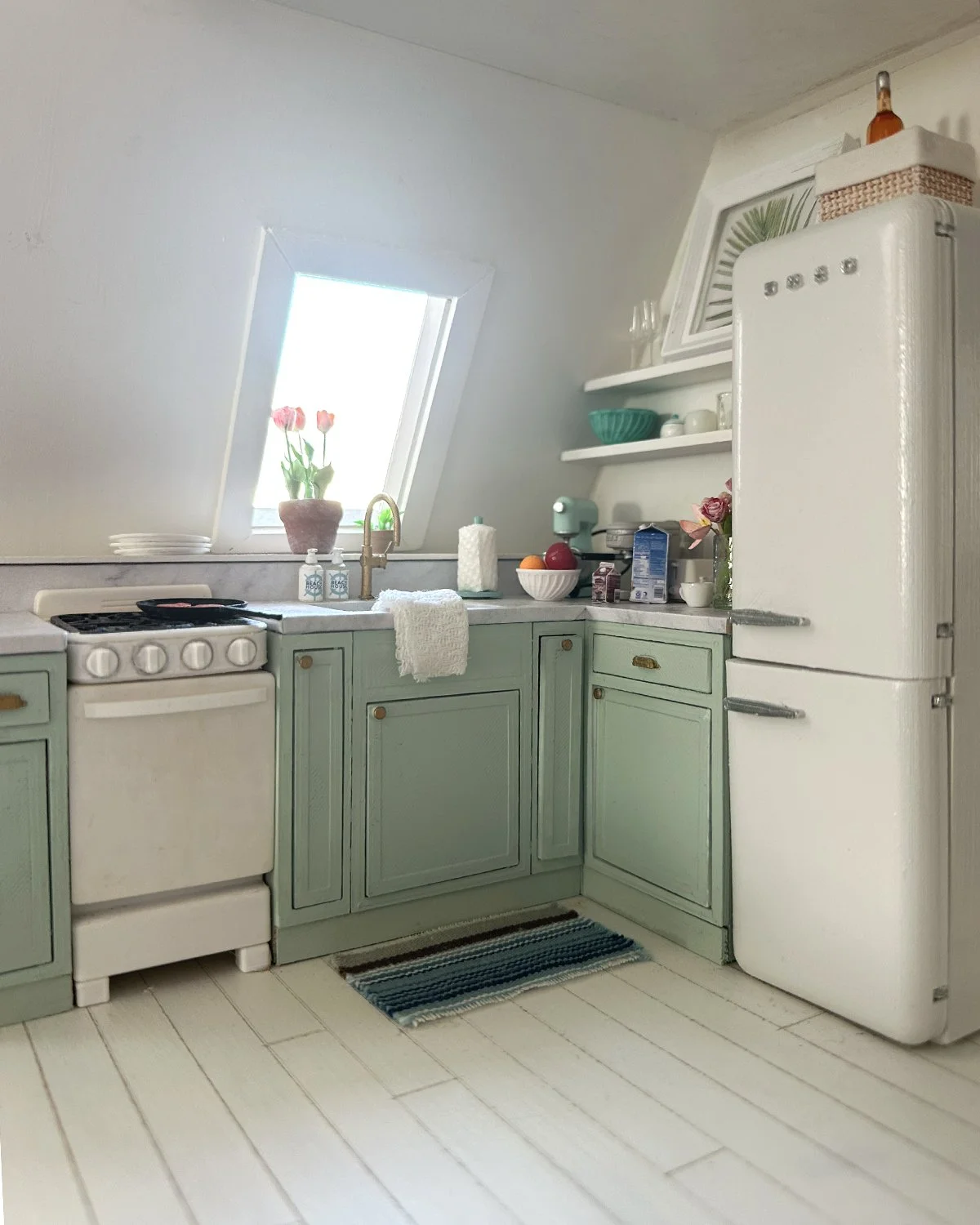 A cozy miniature vintage kitchen with pastel green cabinets, a white refrigerator, and a small window with pink tulips in a pot. The countertop holds various kitchen items, and there is a small striped rug on the light wooden floor.