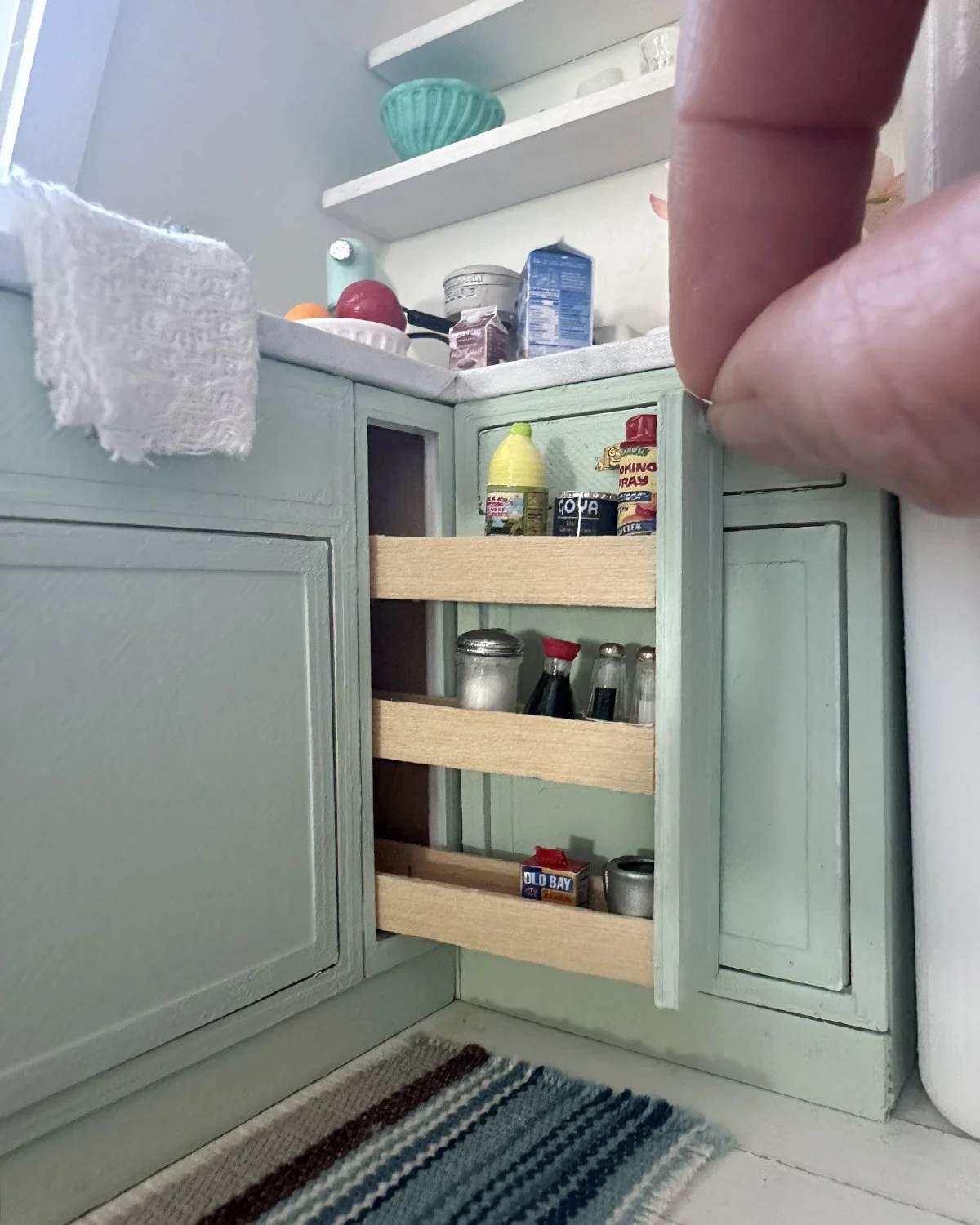 Close-up of a miniature kitchen cabinet with open wooden shelves containing small bottles and cans, next to a countertop with food items and a towel hanging on the side.
