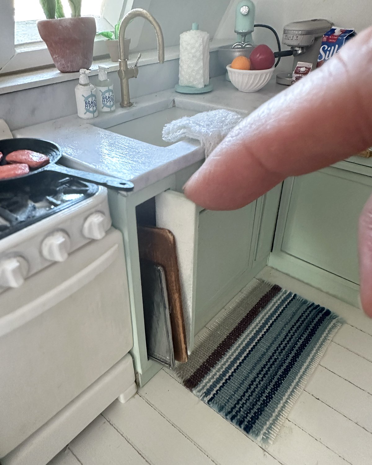 Close-up of a kitchen scene with a person's arm reaching over a countertop. The stove has a frying pan with raw sausages. A sink and faucet are in the background, with dish soap and paper towels on the counter. A woven rug is on the floor, and variou