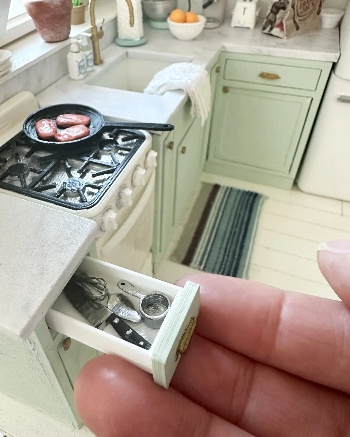 Miniature kitchen scene with a small stove, green cabinets, and a miniature drawer with tiny kitchen utensils including a whisk, a spatula, and a measuring spoon. The stove has a cast iron pan with miniature steaks.
