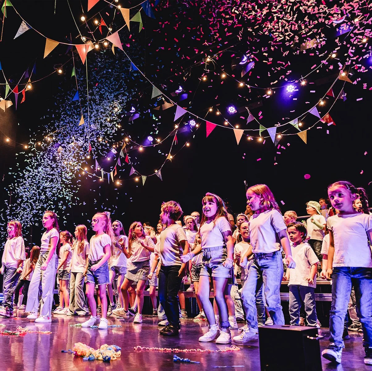 Children performing on stage at a celebration or event, with colorful bunting, string lights, and confetti falling from above.