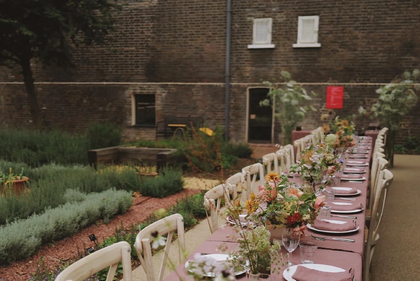 Outdoor garden dining setup with a long table, chairs, and floral centerpieces near a brick building.