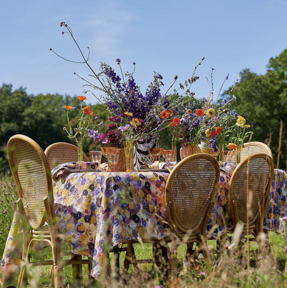 Outdoor table setting with a floral tablecloth, wicker chairs, and vibrant flowers in vases against a scenic background.