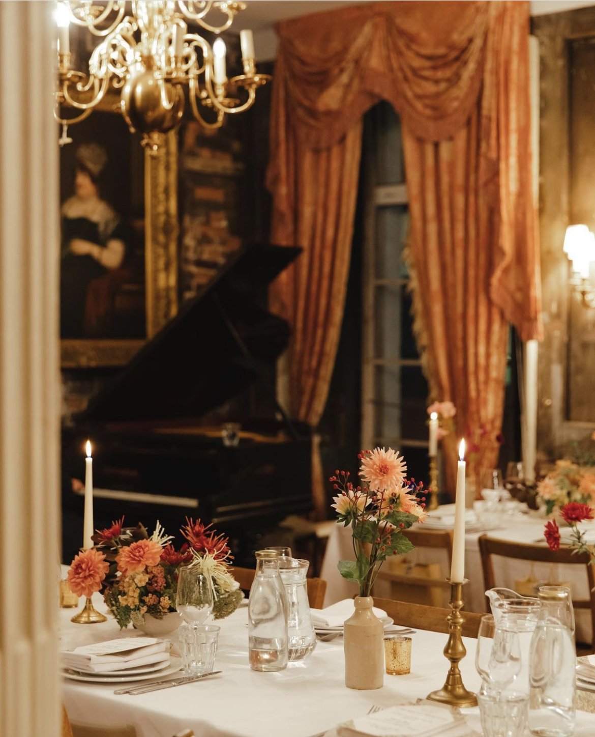 Elegant dining room with candlelit table, flower arrangements, chandelier, and a grand piano in the background.