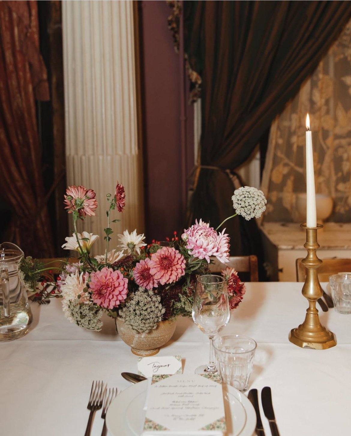 Elegant dining table setup with a floral centerpiece of pink and white flowers, a lit candle, wine and water glasses, cutlery, plates, and a menu card.