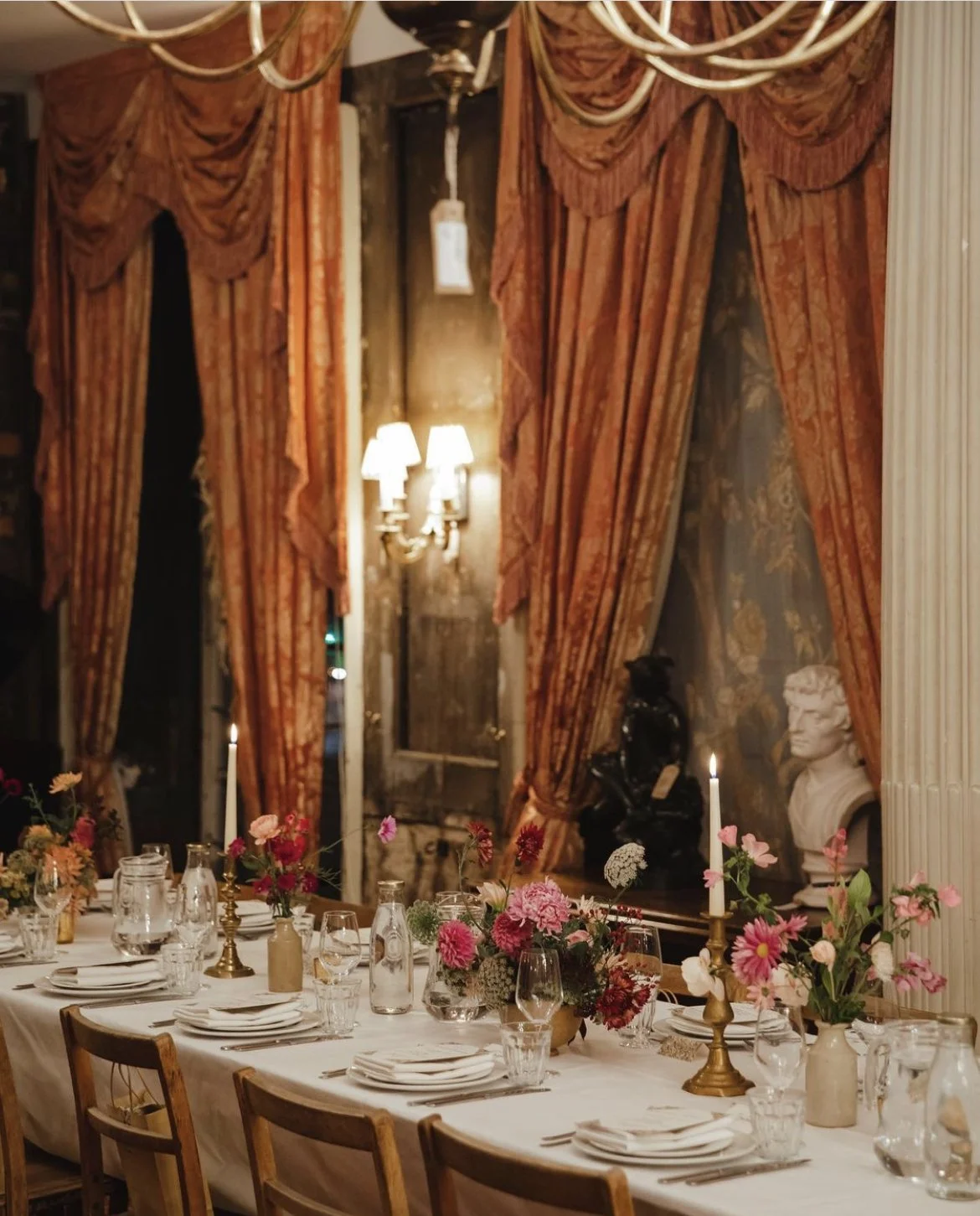 Elegant dining table setting with floral centerpieces, candles, and vintage decor in a dimly lit room with draped curtains.